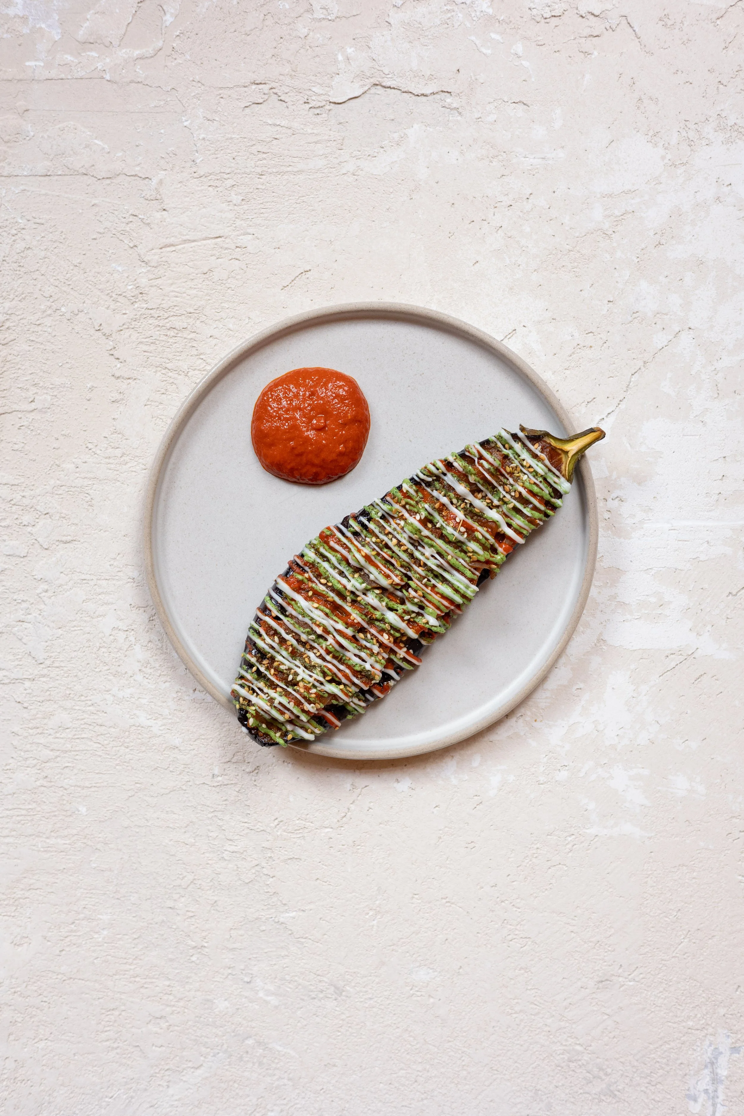 A plate with an eggplant dish topped with sauce and drizzled with sauces, served with a side of red sauce.