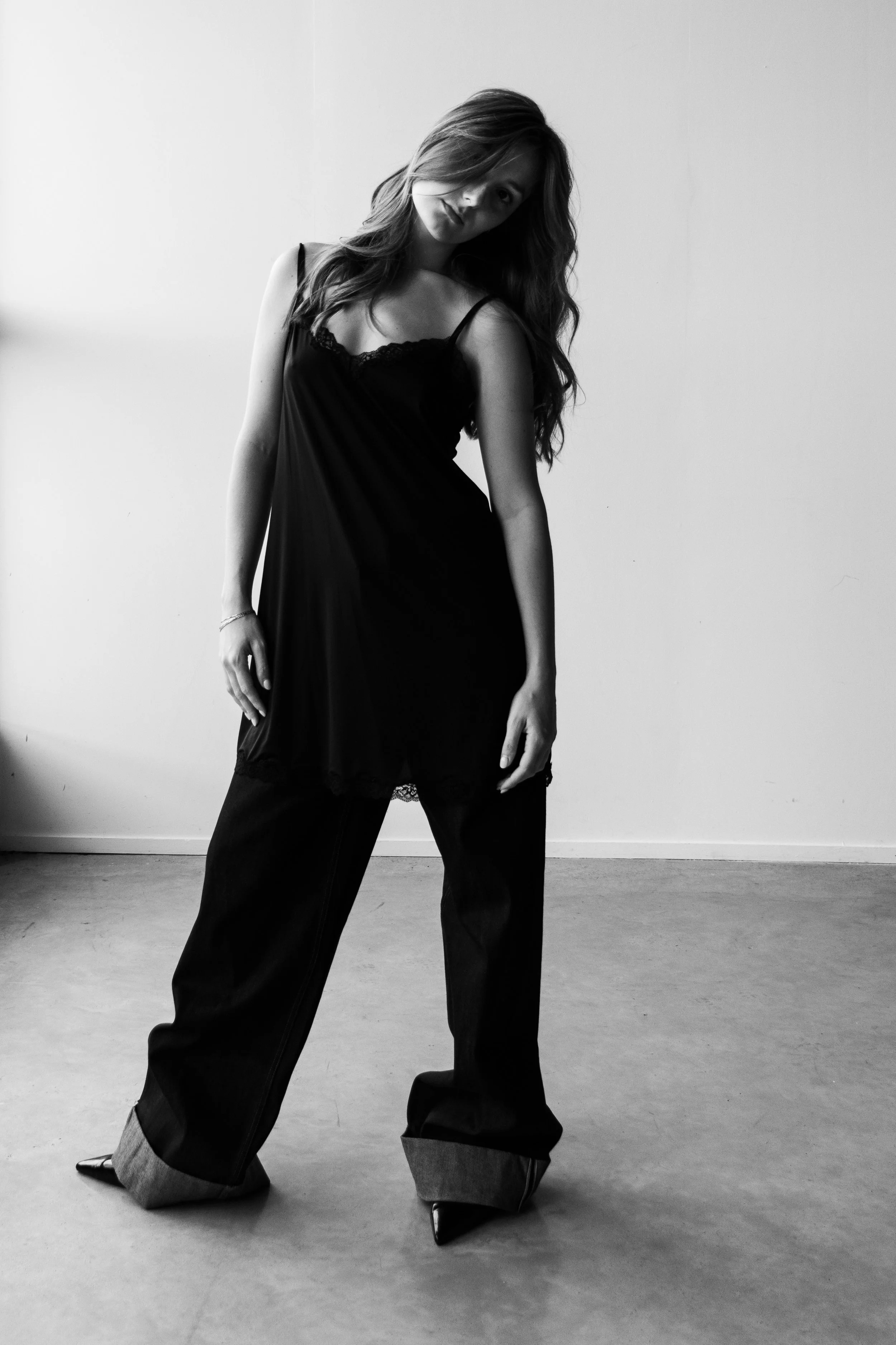 A woman in a black dress and wide-legged jeans with pointed shoes standing in a room with a plain wall and floor, black and white photograph.