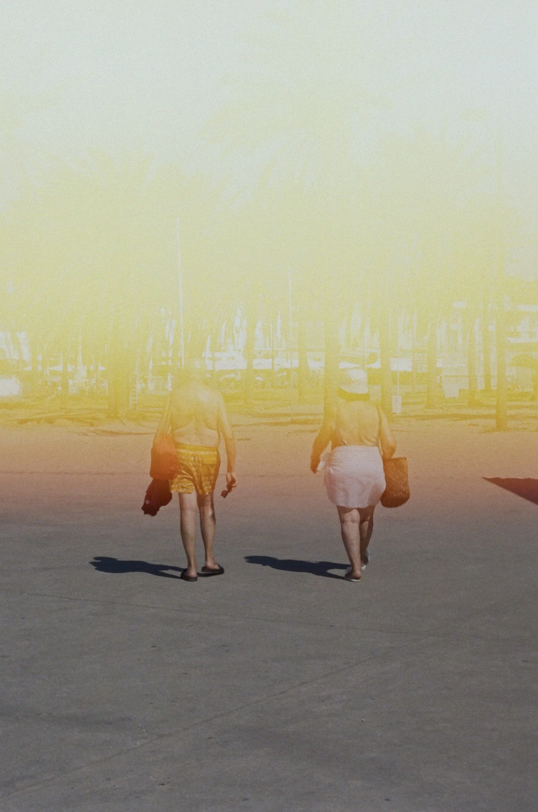Two women walking on a beach in summer, carrying bags and wearing hats, with yellow light overlay.
