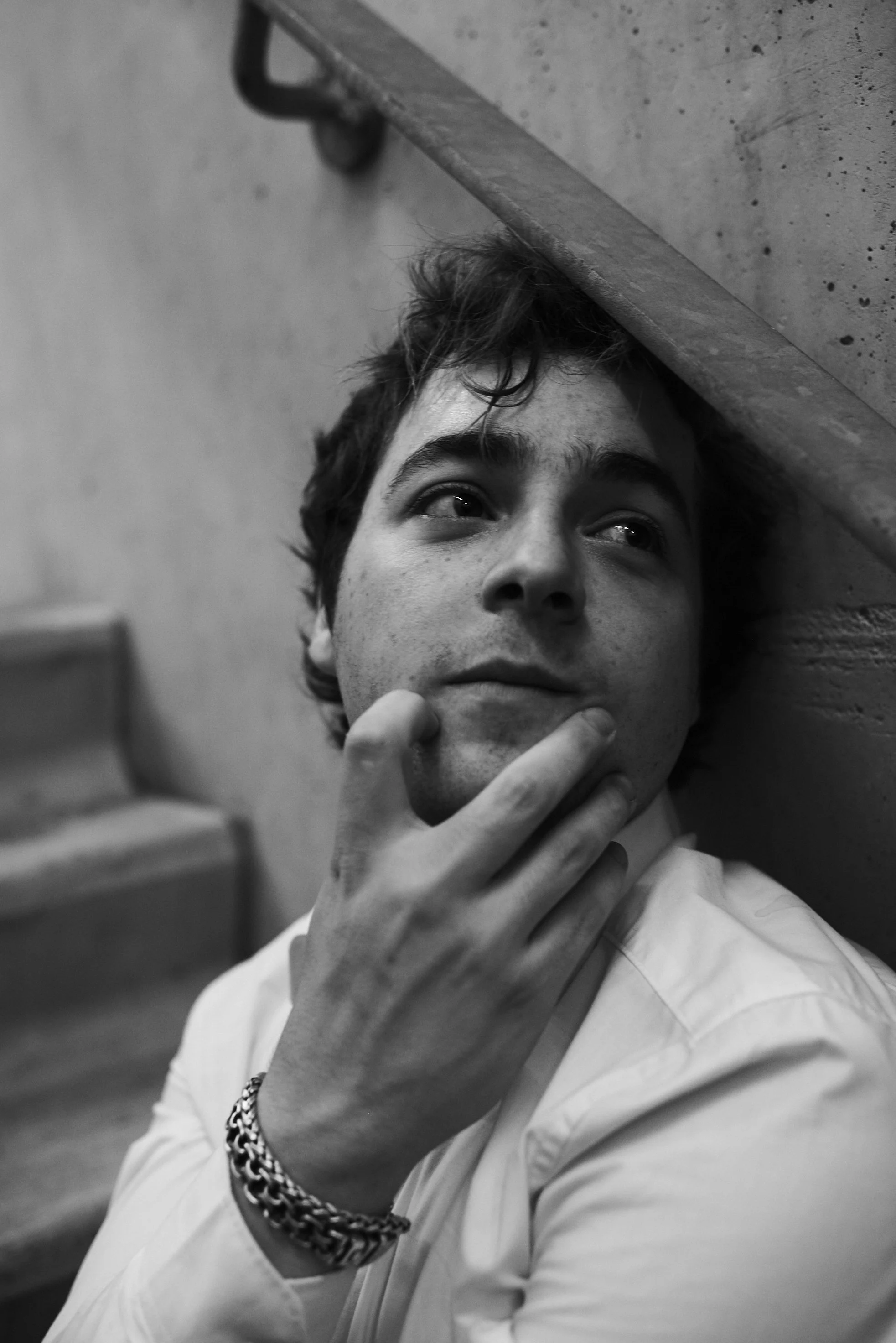 A young man with curly hair, wearing a white shirt and a bracelet, sitting near stairs and leaning against a wall, gazing thoughtfully to the side with his hand on his chin.