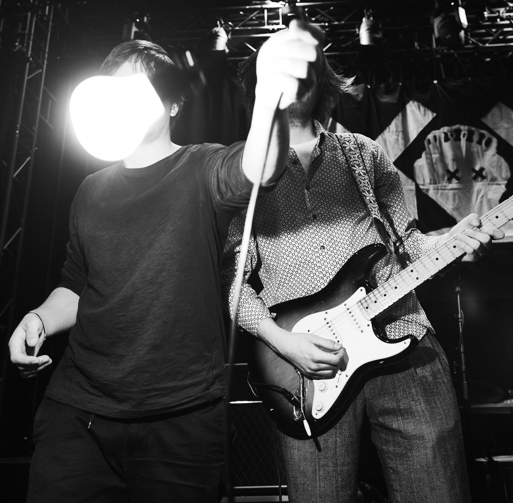 Two musicians on stage, one holding a microphone and the other playing an electric guitar, with a skull and crossbones banner in the background.