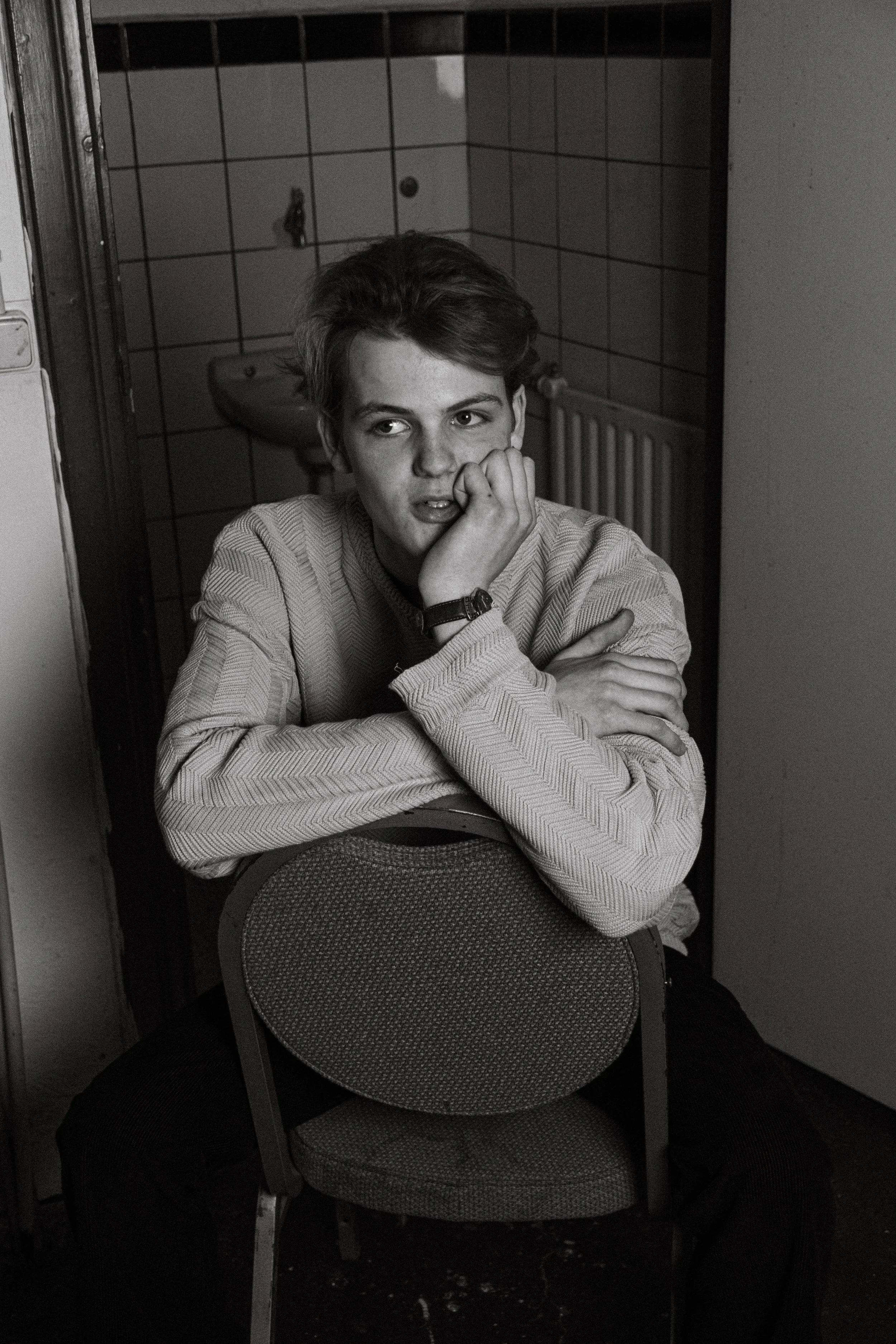 A young man with short hair sitting backwards on a chair, resting his head on his hand, looking to the side in a room with tiled walls and a radiator in the background.