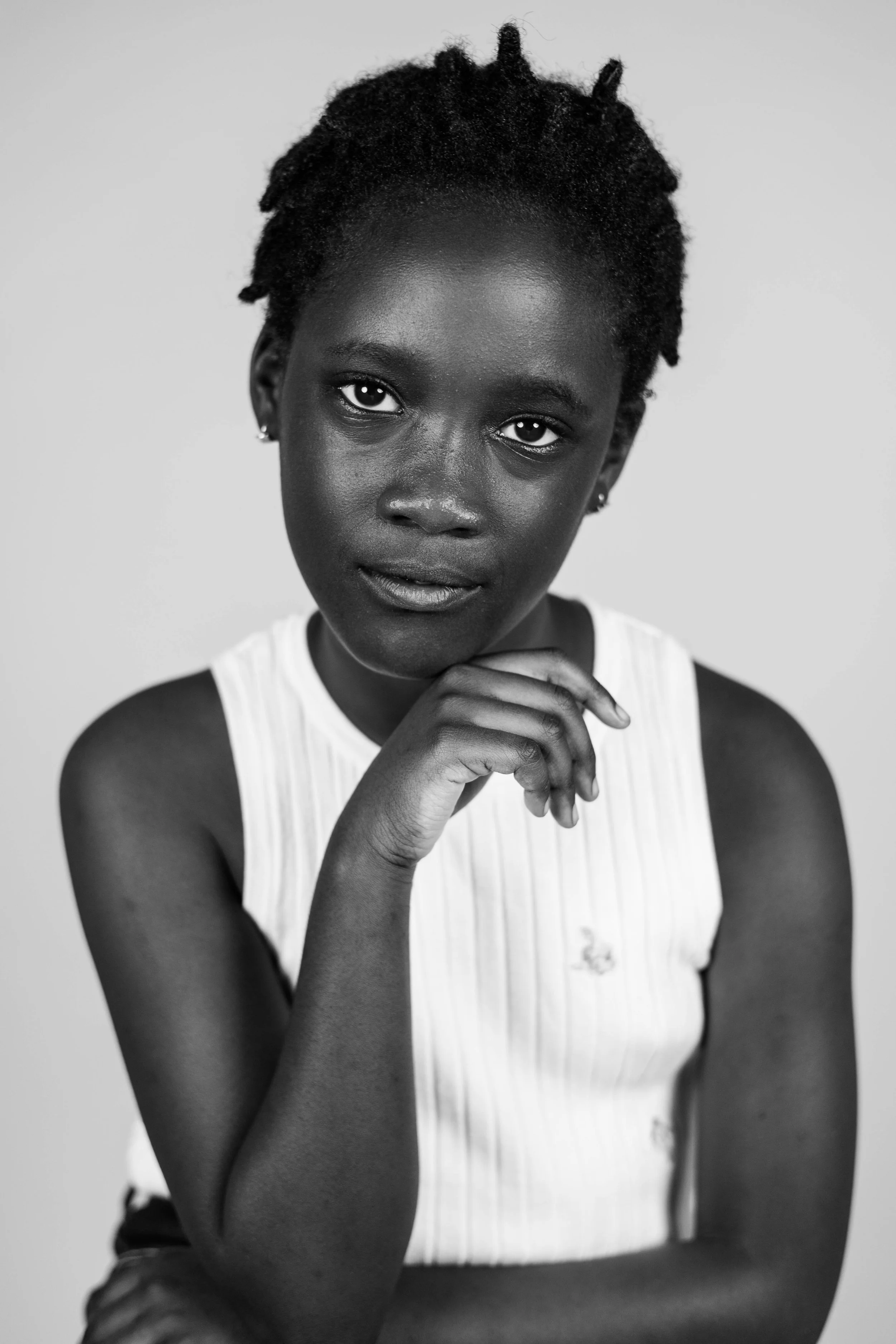 Black and white portrait of a young woman with short natural hair, wearing a sleeveless top, touching her chin with her right hand, looking directly at the camera.