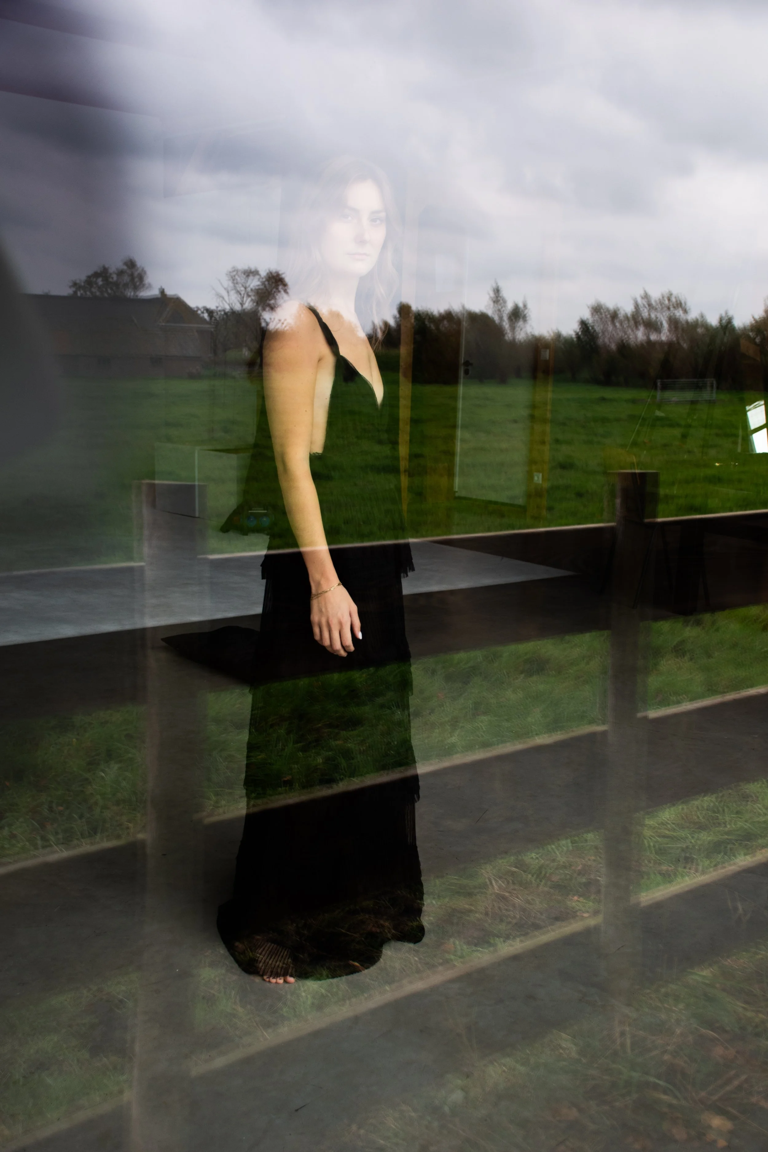 A woman dressed in black standing outdoors, seen through a glass window that reflects the surrounding landscape of grass, trees, and cloudy sky.