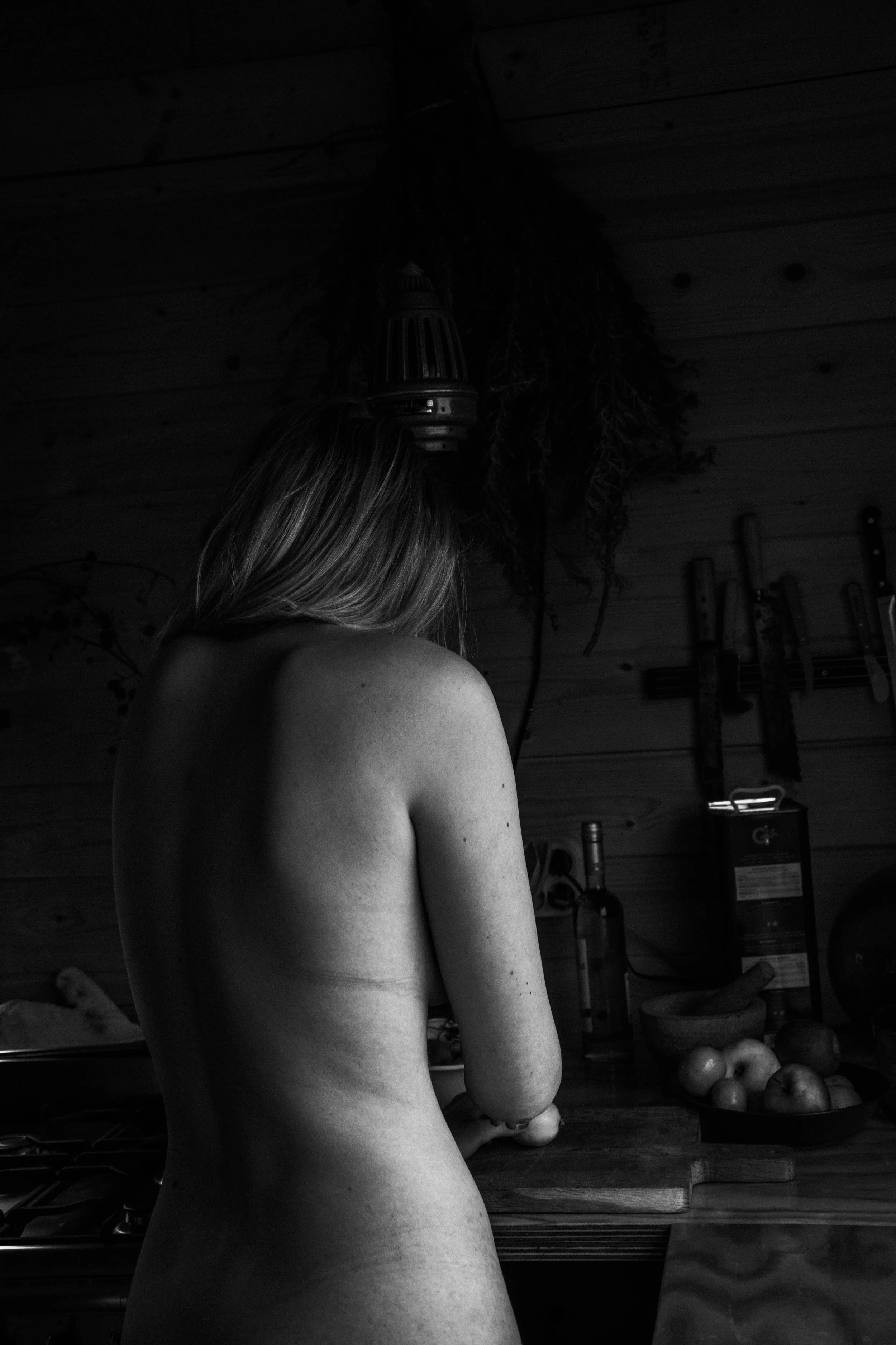 A woman with long hair, topless, seen from behind, standing in a rustic kitchen, preparing food with a cutting board and vegetables.