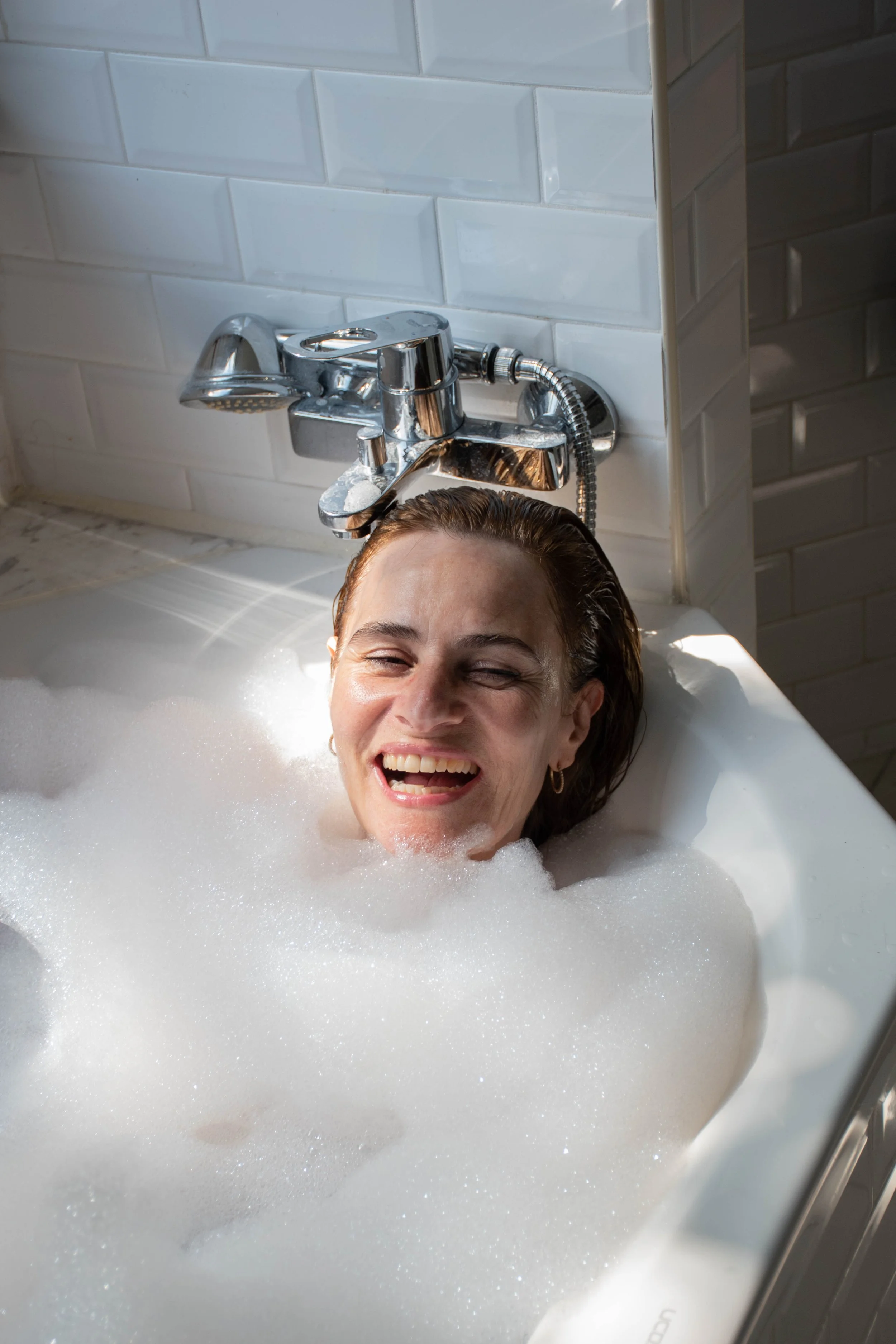 A woman with wet hair smiling and laughing while taking a bath in a bathtub filled with soap bubbles, with a tiled wall and a chrome faucet above her.