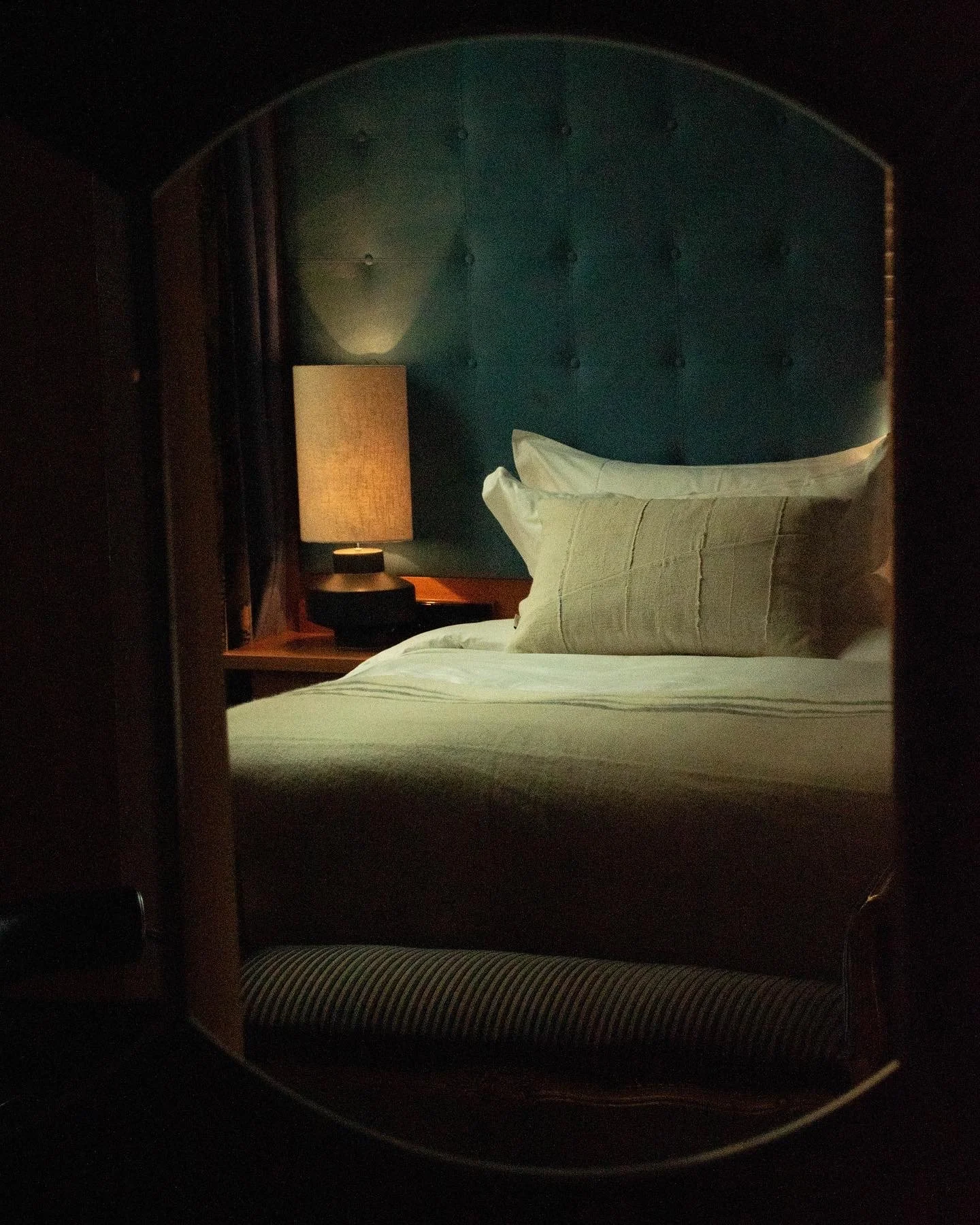 A cozy bedroom with a bed, pillows, and a nightstand with a lamp, viewed through a mirror.