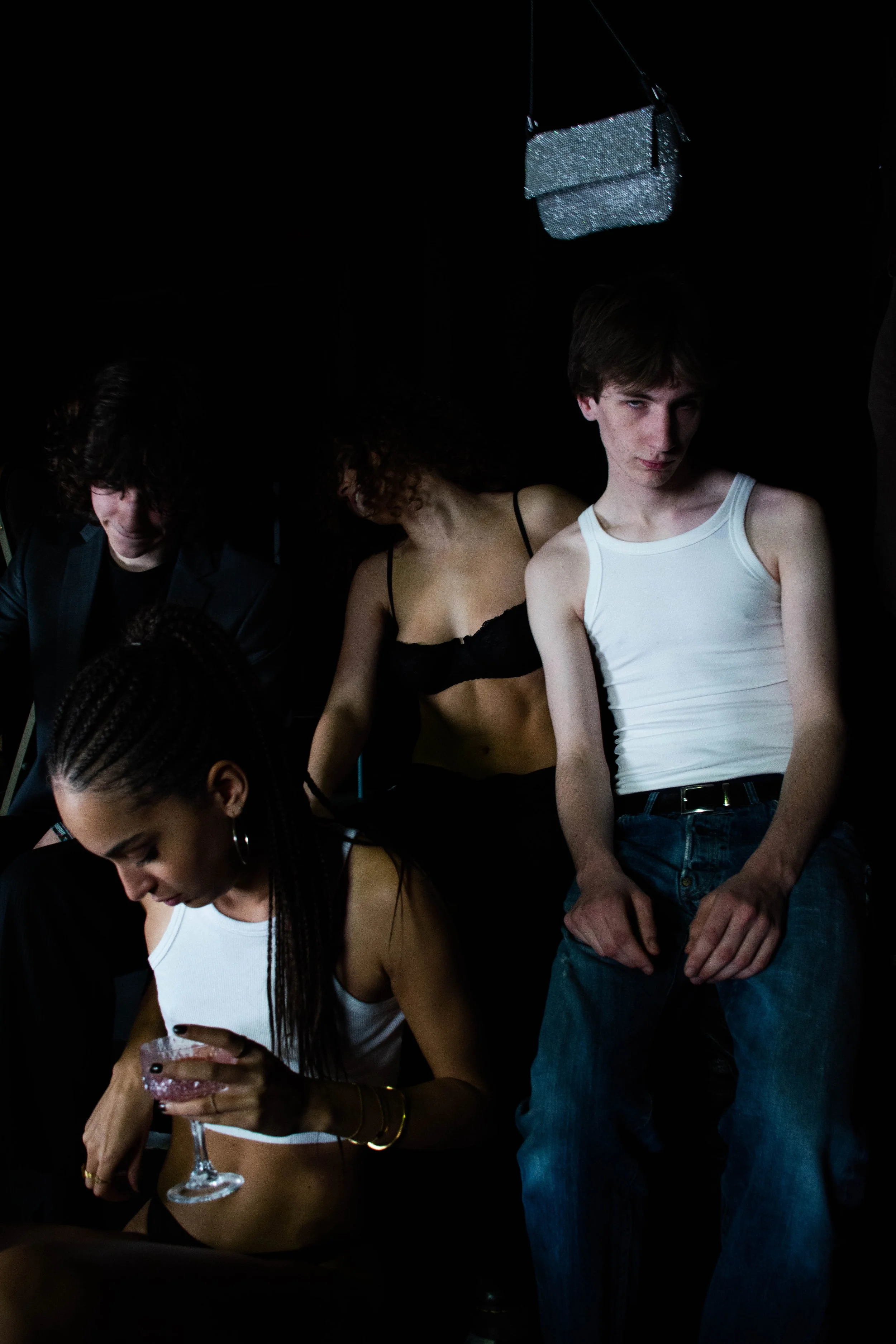 Group of young people in a dimly lit setting, with one person holding a drink, others dressed casually, some in tank tops or lingerie, with a dark background and a small ceiling light.