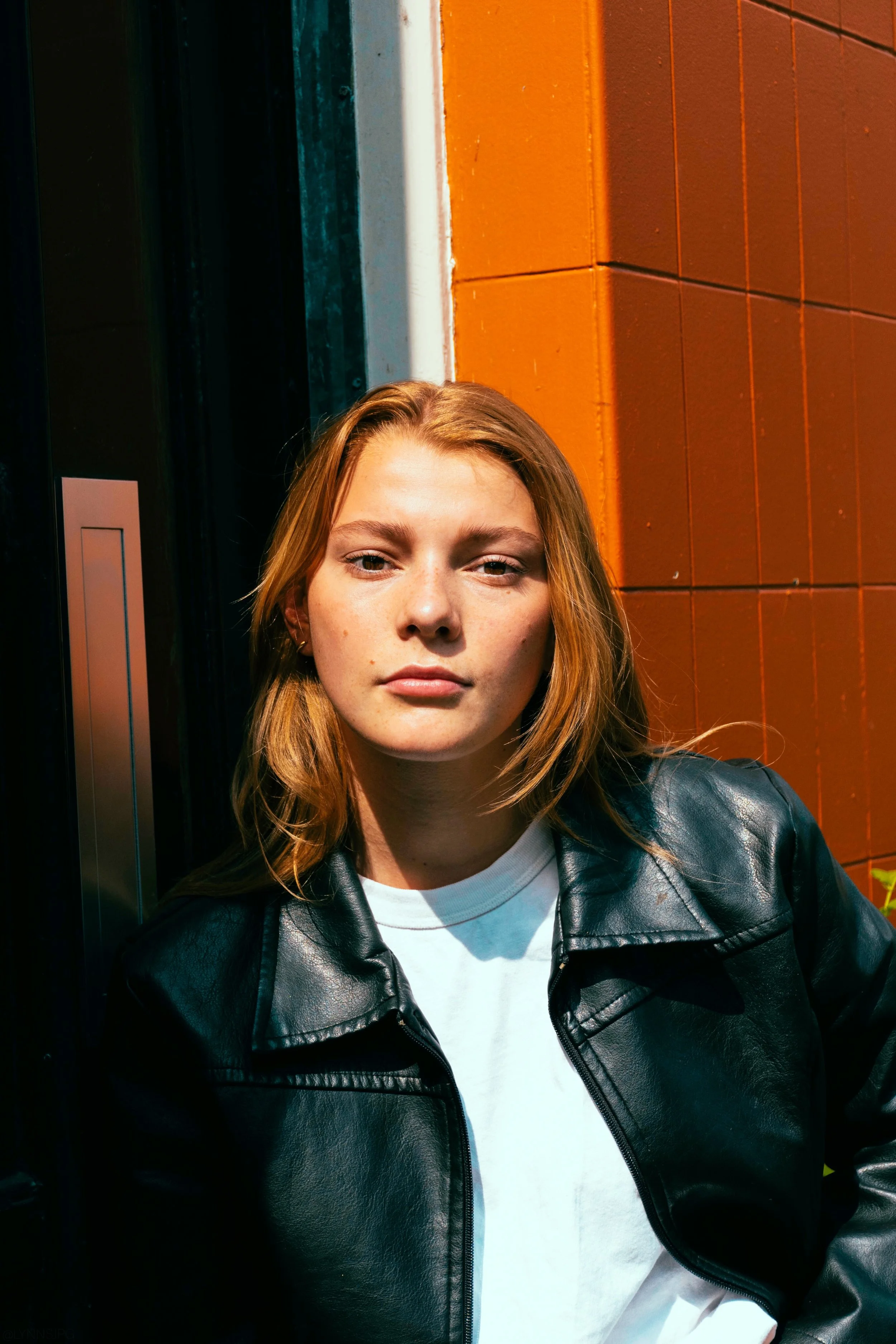 A young woman with red hair, wearing a black leather jacket and white T-shirt, leaning against a wall with orange and black panels, in bright sunlight.