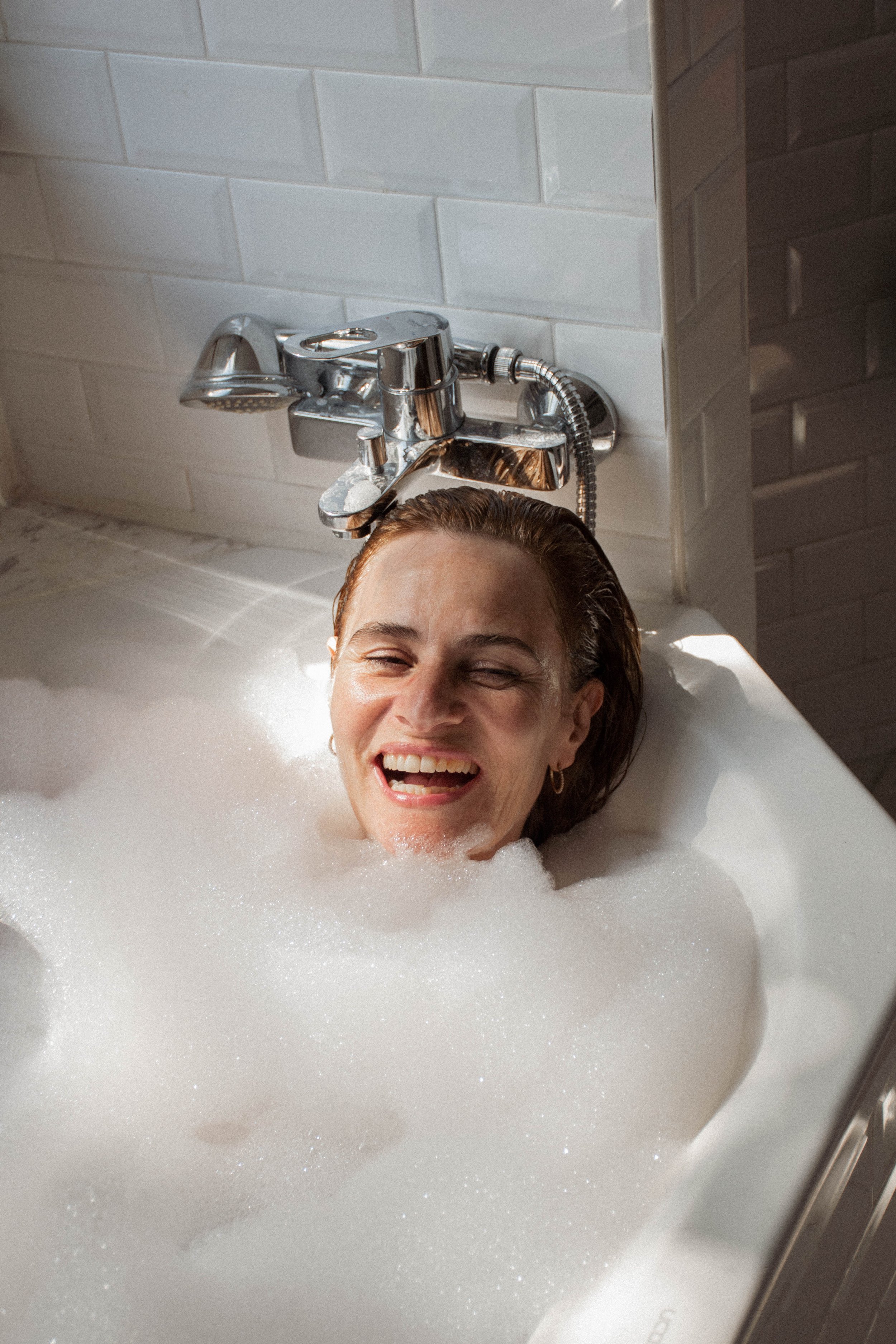 A woman with reddish hair enjoying a bubble bath in a white bathtub with bubbles, smiling with her eyes closed.