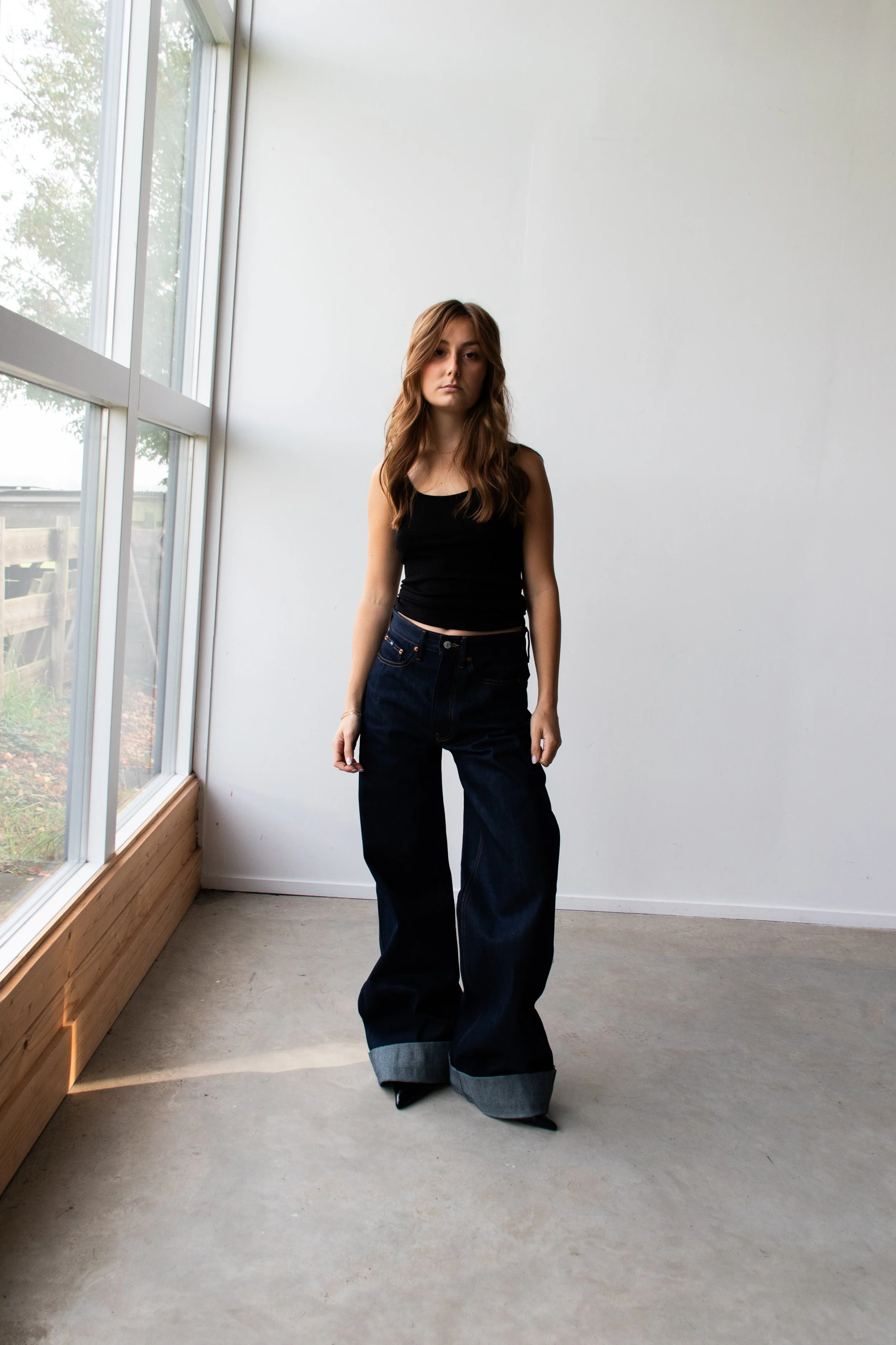 A young woman with long wavy hair stands inside a room near a large window, wearing a black tank top and wide-leg dark jeans.