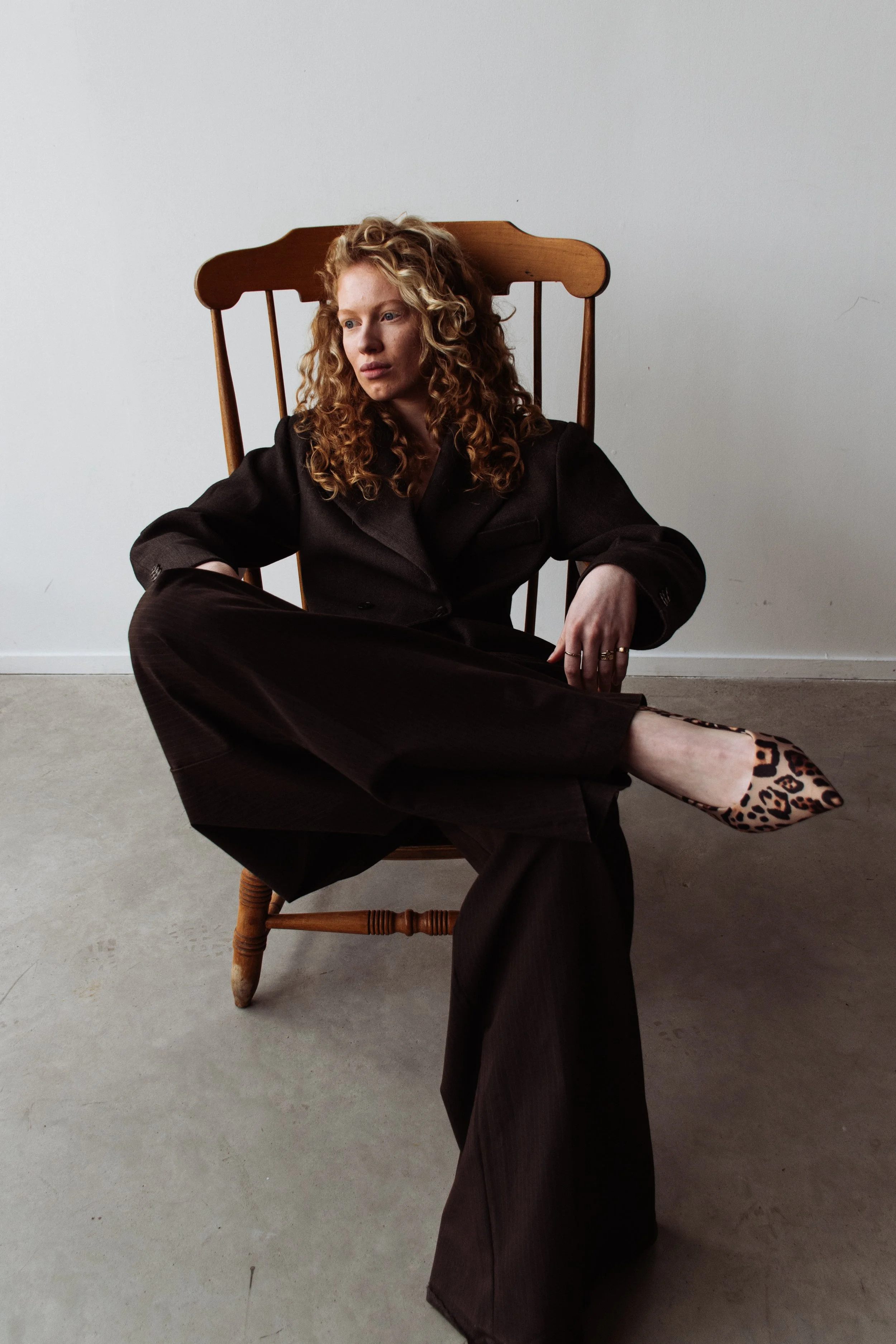 Woman with curly red hair sitting in a wooden chair, wearing a dark blazer, wide-leg pants, and leopard-print shoes, against a plain white wall.