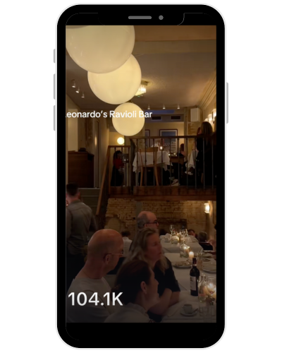 A group of people dining at Leonardo's Ravioli Bar, sitting at tables with candles, in a warmly lit restaurant with hanging spherical lights and a second-floor balcony.
