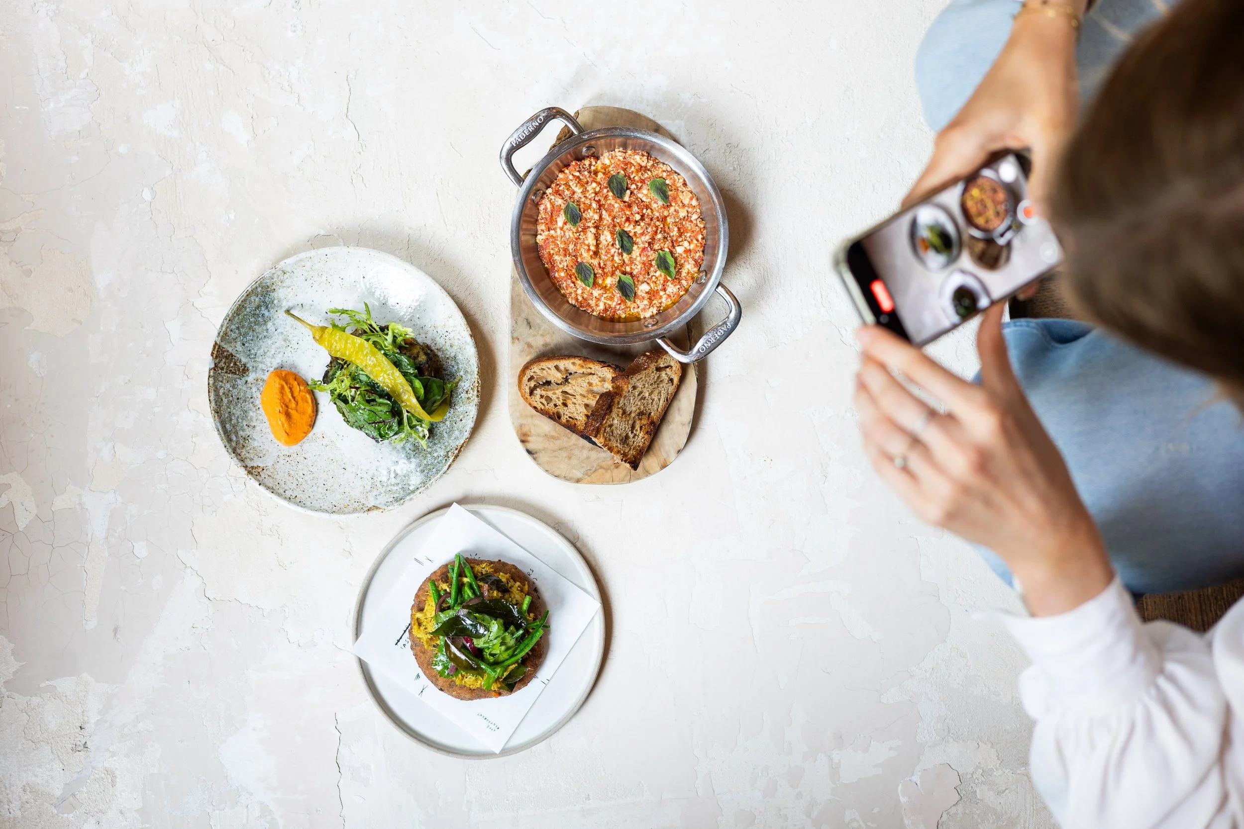 Top-down view of a person taking a photo of three plated dishes and a pot of food on a textured white surface.