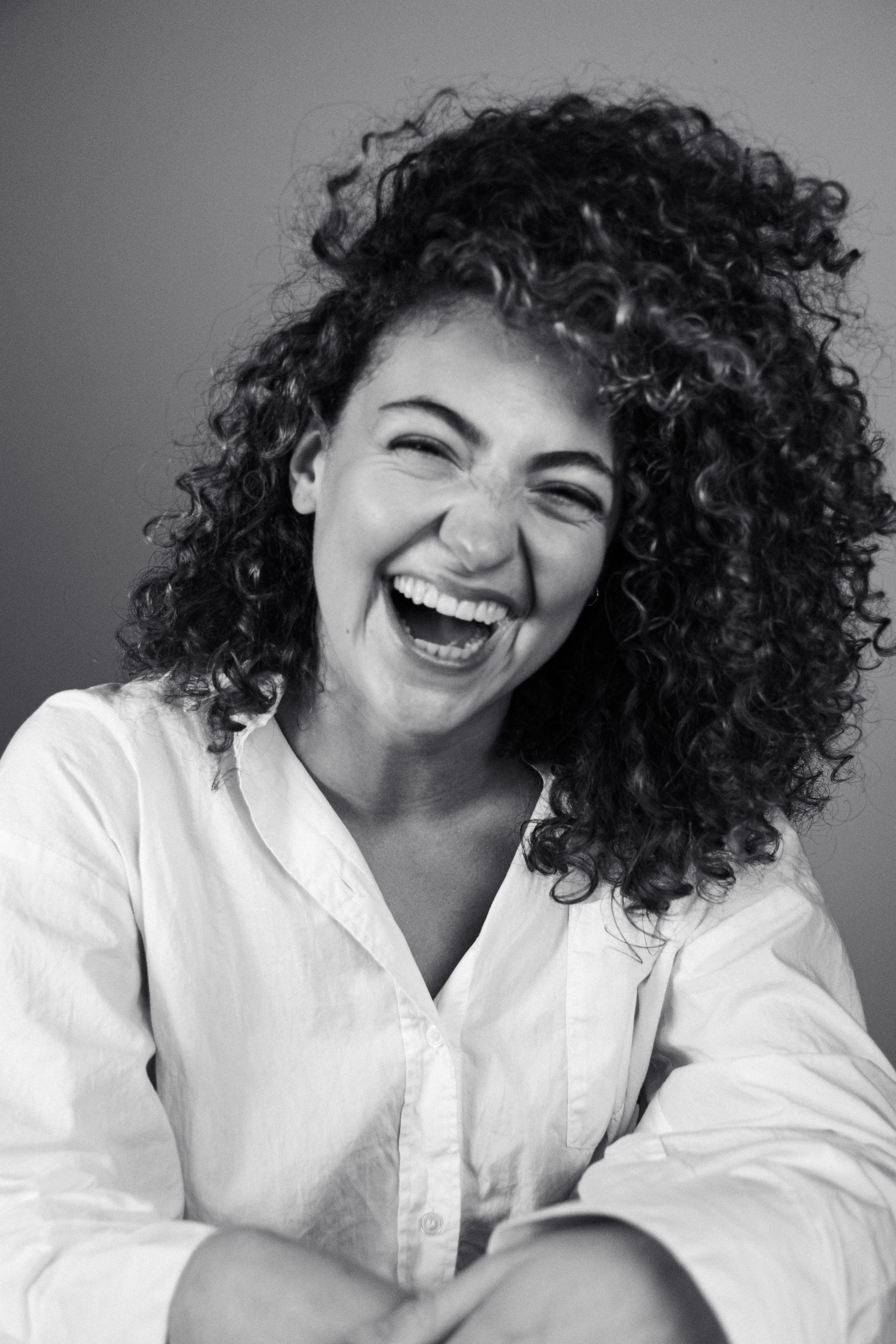 A woman with curly hair laughing, wearing a light-colored shirt, in a black and white photo.