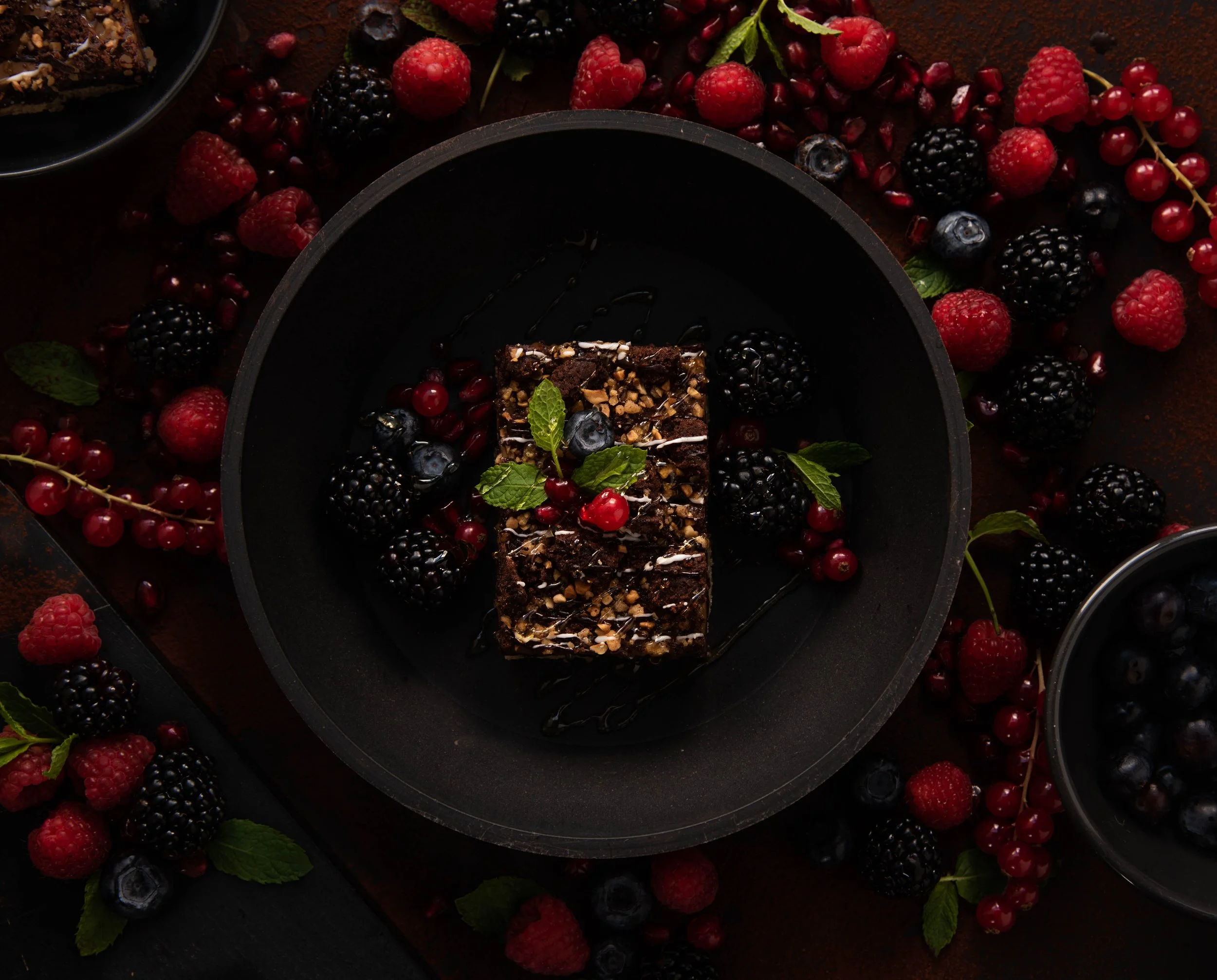 Chocolate dessert squares garnished with mint and berries on a black plate, surrounded by scattered red and black raspberries, blackberries, and red currants.
