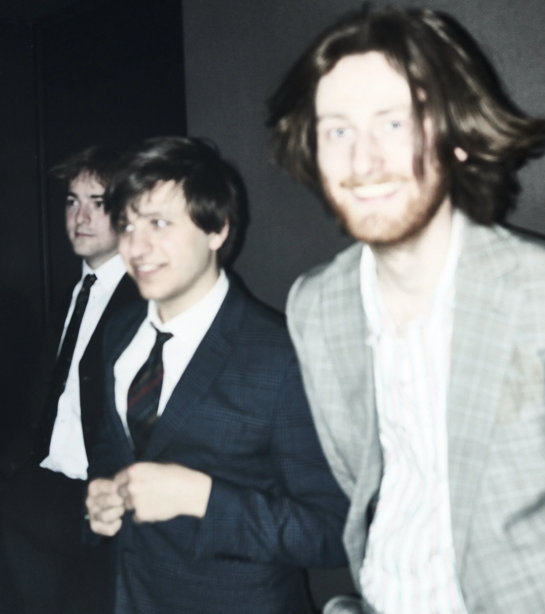 Three young men dressed in suits and ties standing side by side, smiling, against a dark background.