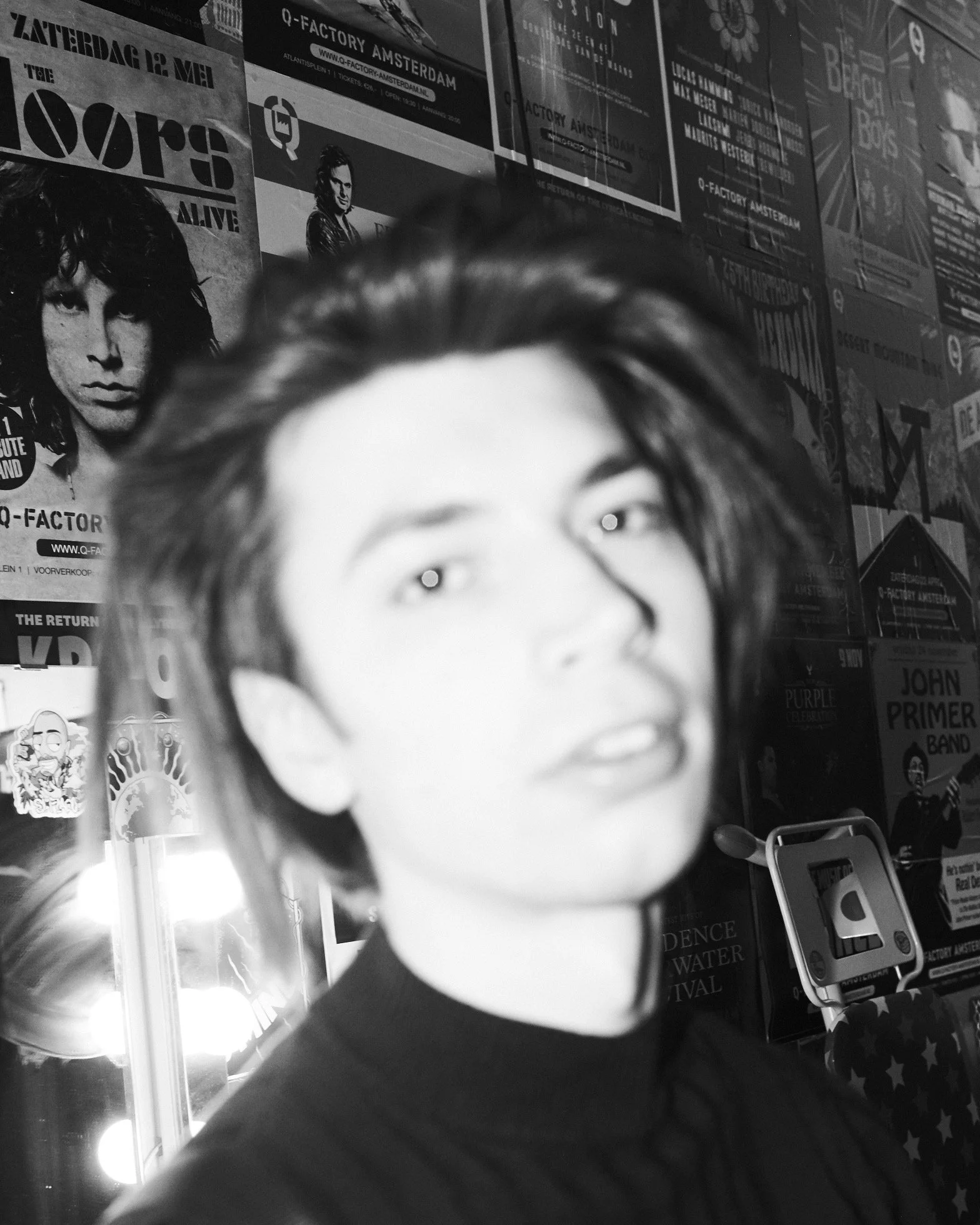 A person with short hair takes a black and white selfie in front of a wall covered with posters and flyers for concerts and events.