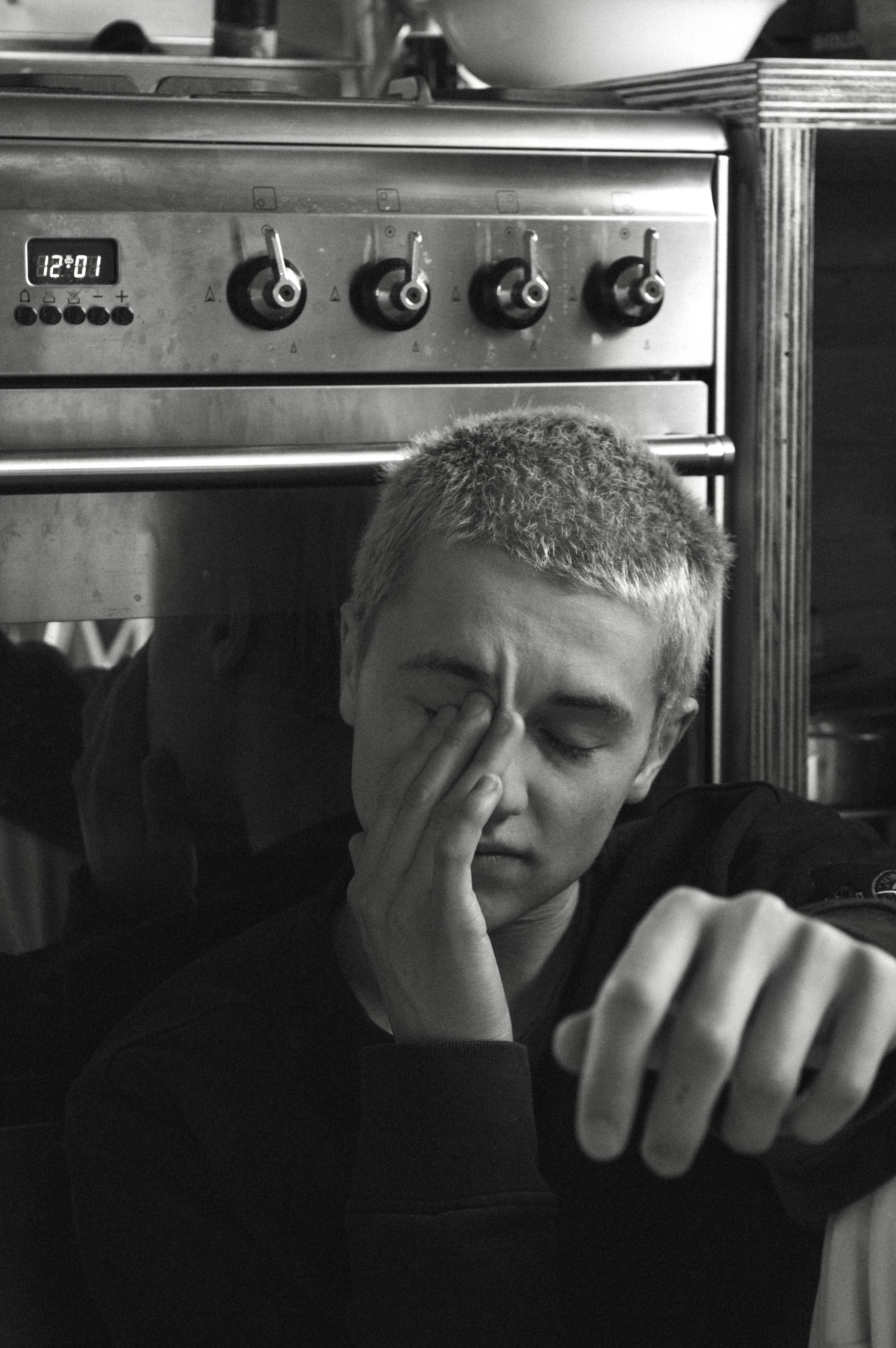 A young man with short hair, sitting on the floor with a distressed expression, holding his face with one hand, and looking at his watch. Behind him is a stainless steel stove with four control knobs and a digital clock showing 12:01.