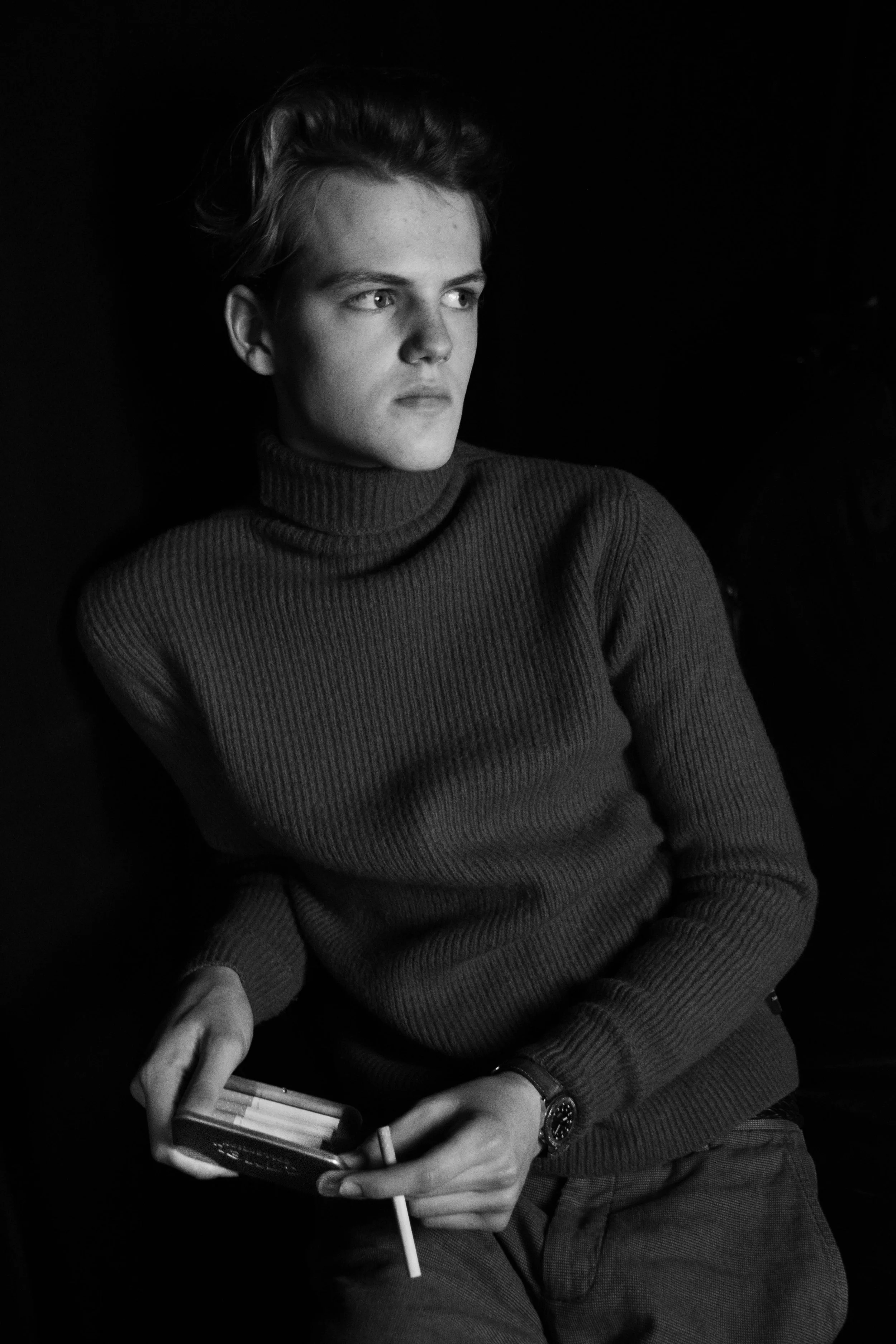 Black and white photo of a young man sitting, wearing a turtleneck sweater, holding a box of cigarettes, with a watch on his left wrist, against a dark background.