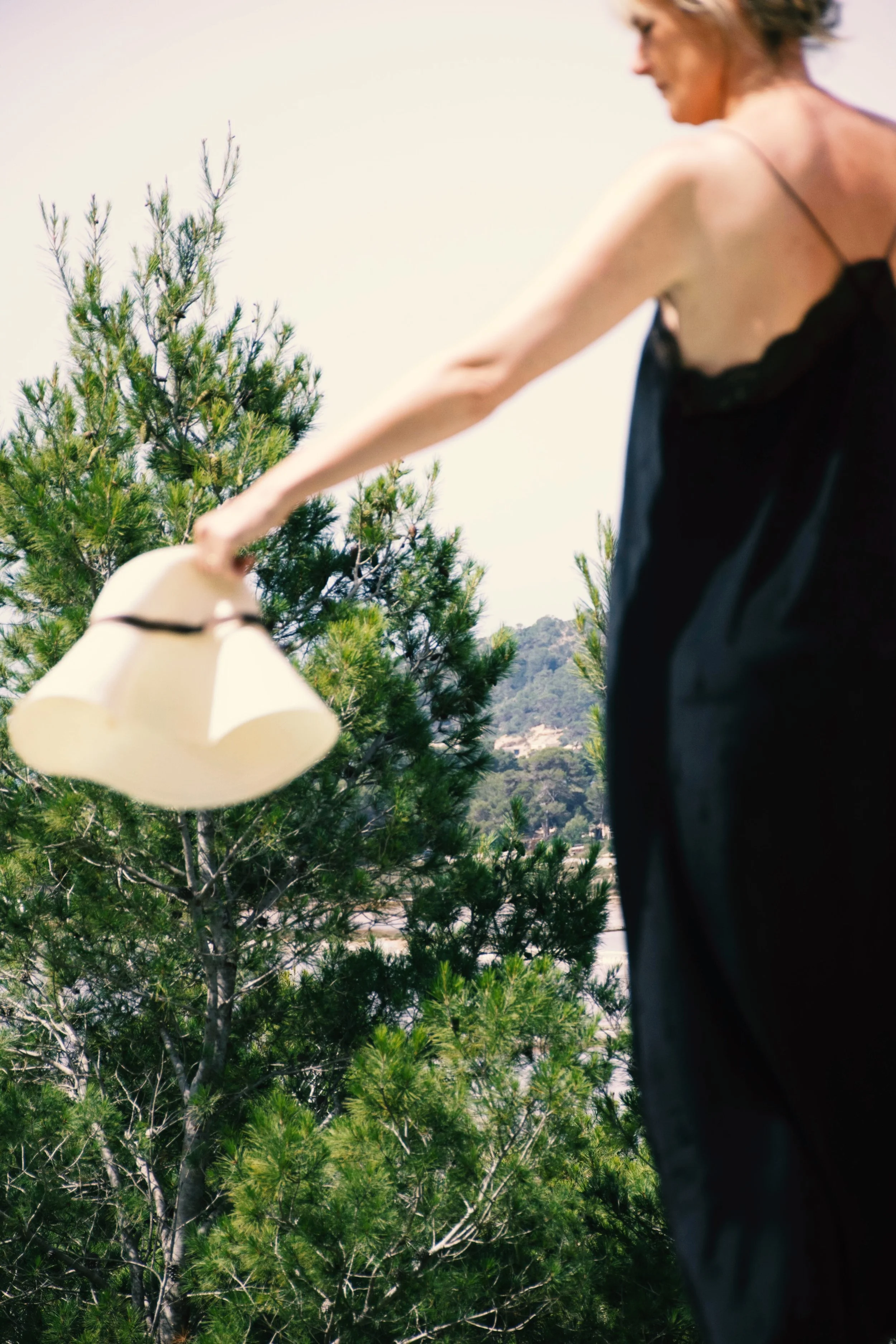 A woman in a black dress holding a beige hat with a black band outside near green trees.