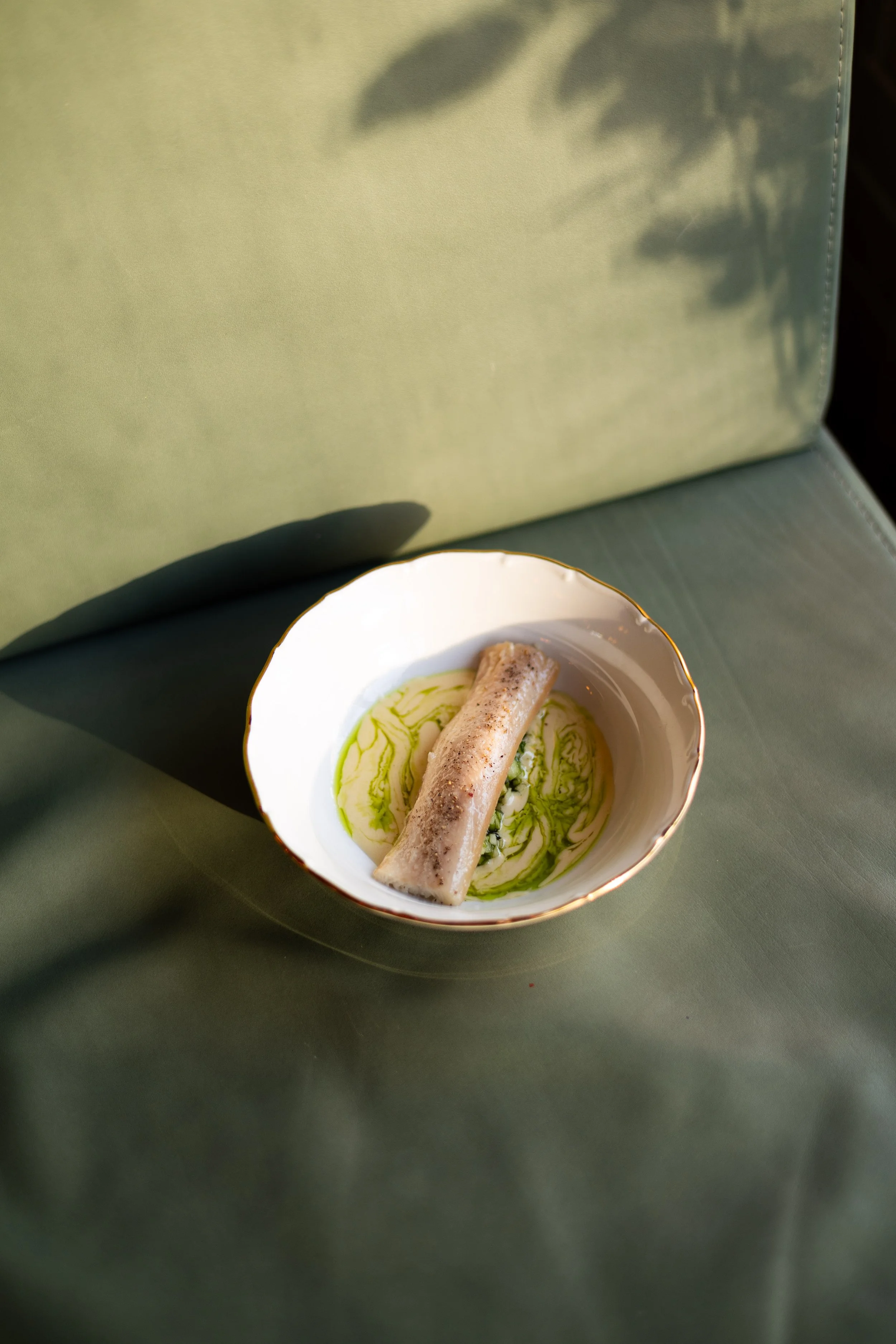 A bowl with a rolled fish fillet on a bed of green sauce, with green liquid swirled around it, set on a green surface.