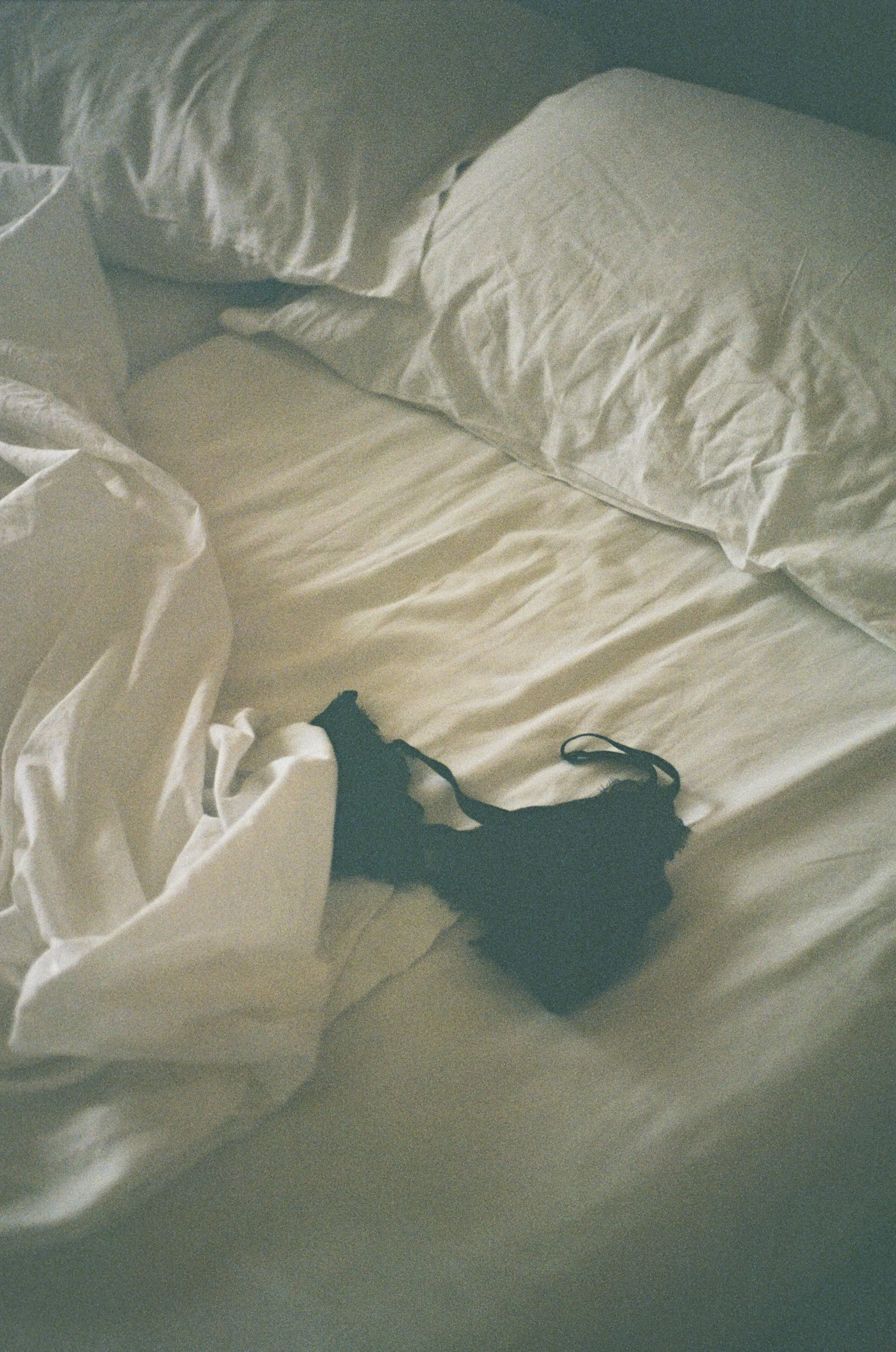 Unmade bed with white sheets, pillows, and a black lingerie piece on the bed.