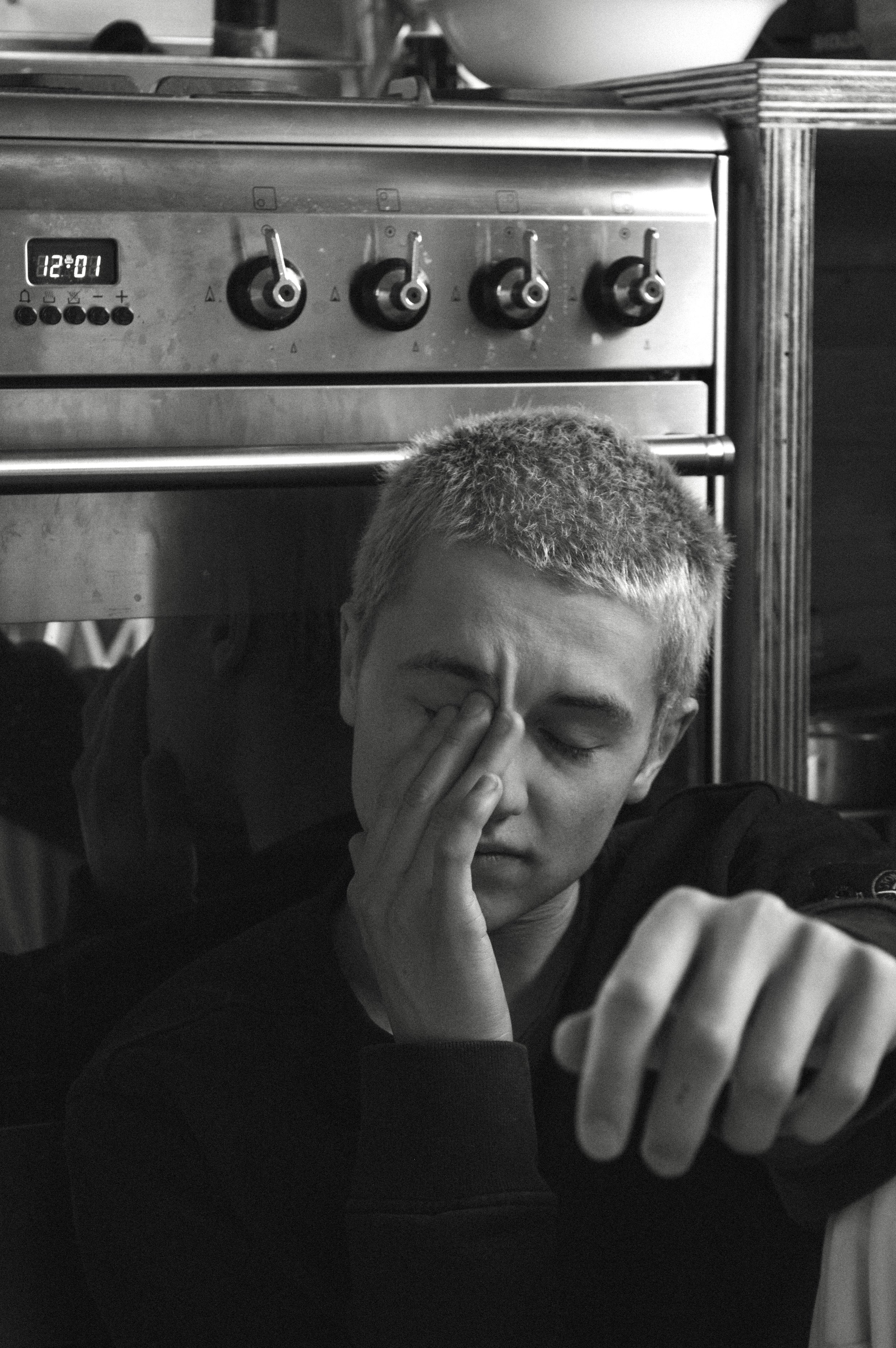 A young person with short hair, resting in front of a kitchen oven, with one eye closed and fingers touching the closed eye, appearing tired or stressed.