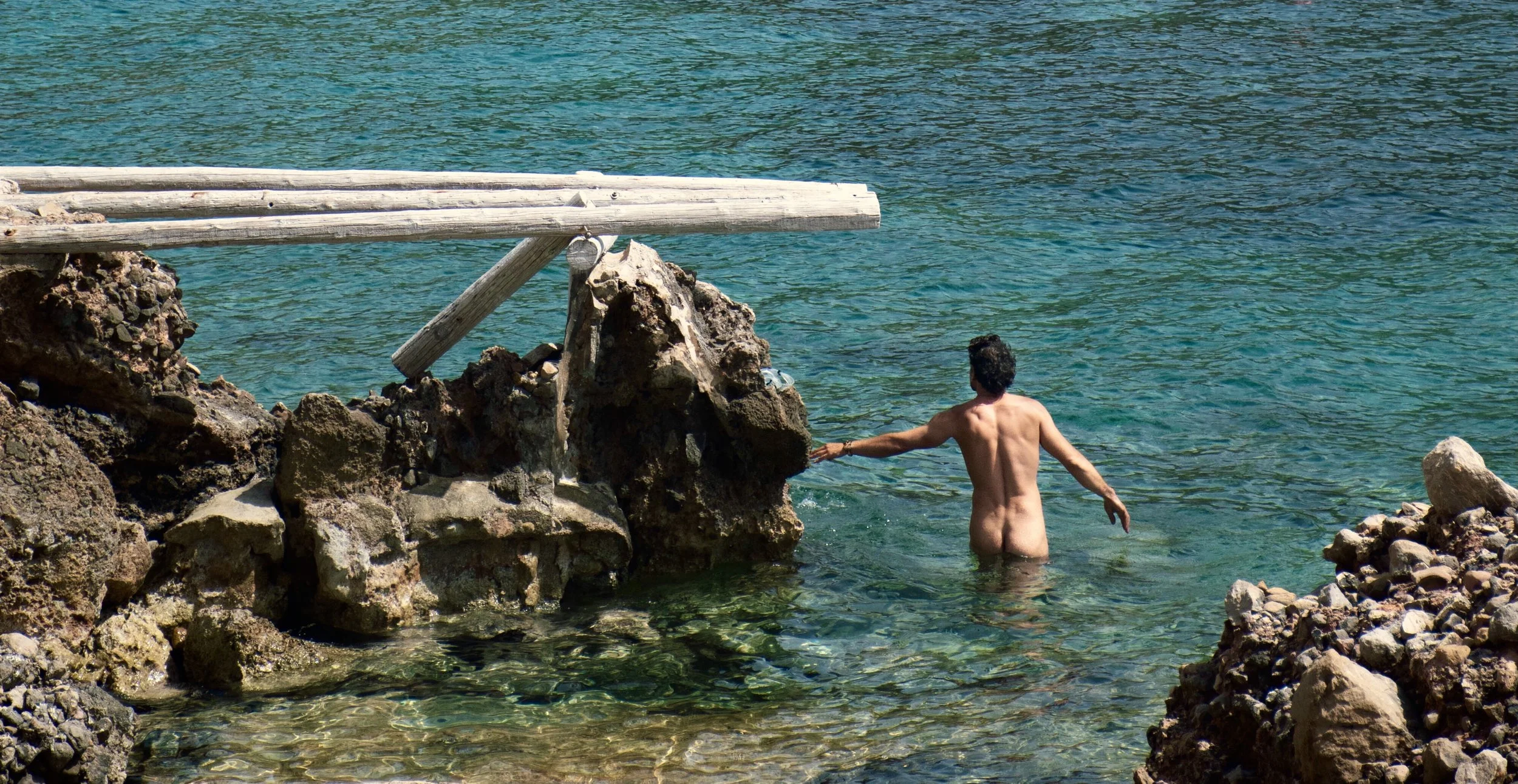 A nude man wading into clear water near rocky shoreline with a wooden pier in the background.