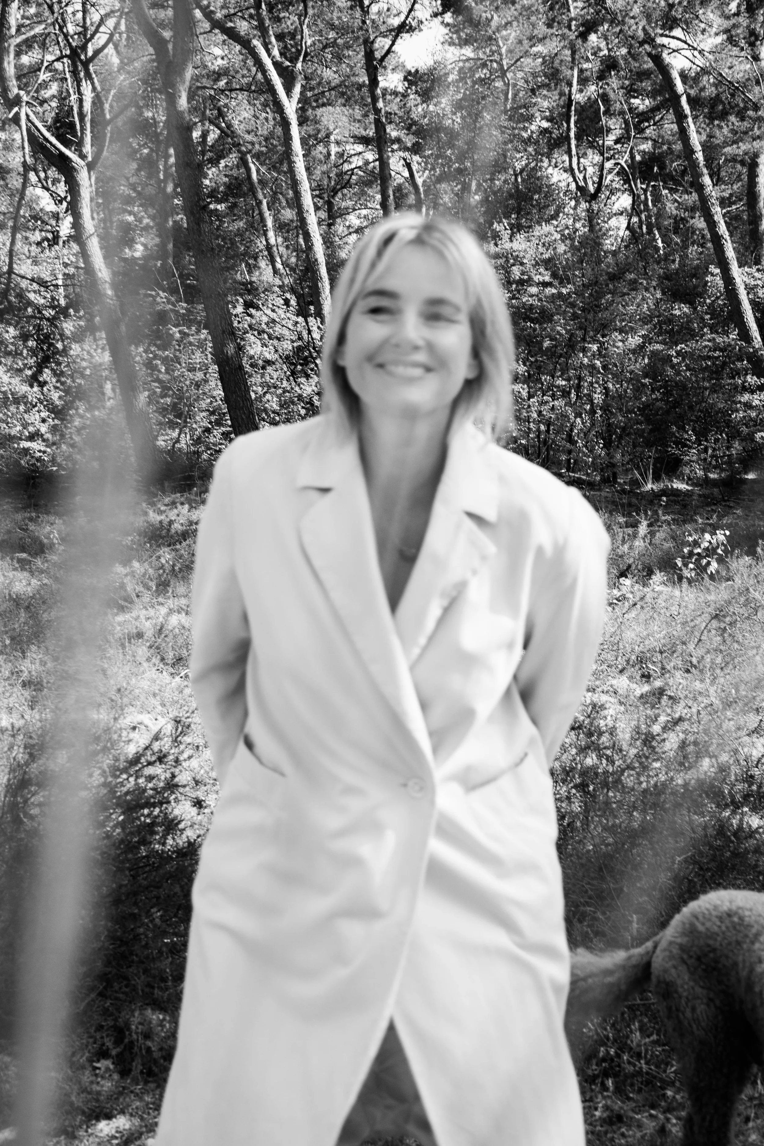 A woman with blonde hair smiling and standing outdoors in a wooded area, wearing a light-colored coat, with trees and foliage in the background.