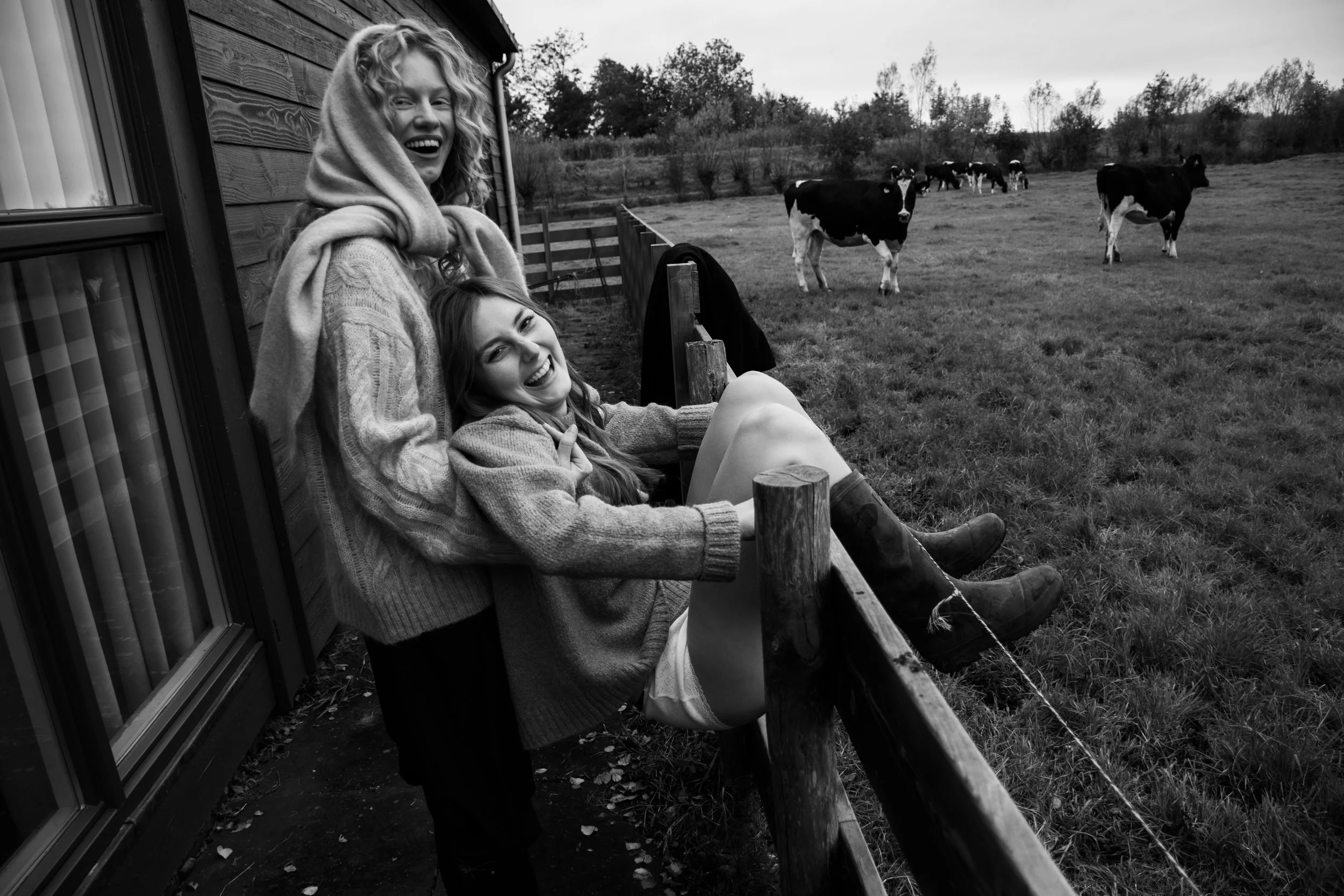 Two women wearing sweaters and boots enjoying themselves on a farm, with cows grazing in the field behind them in a black and white photograph.