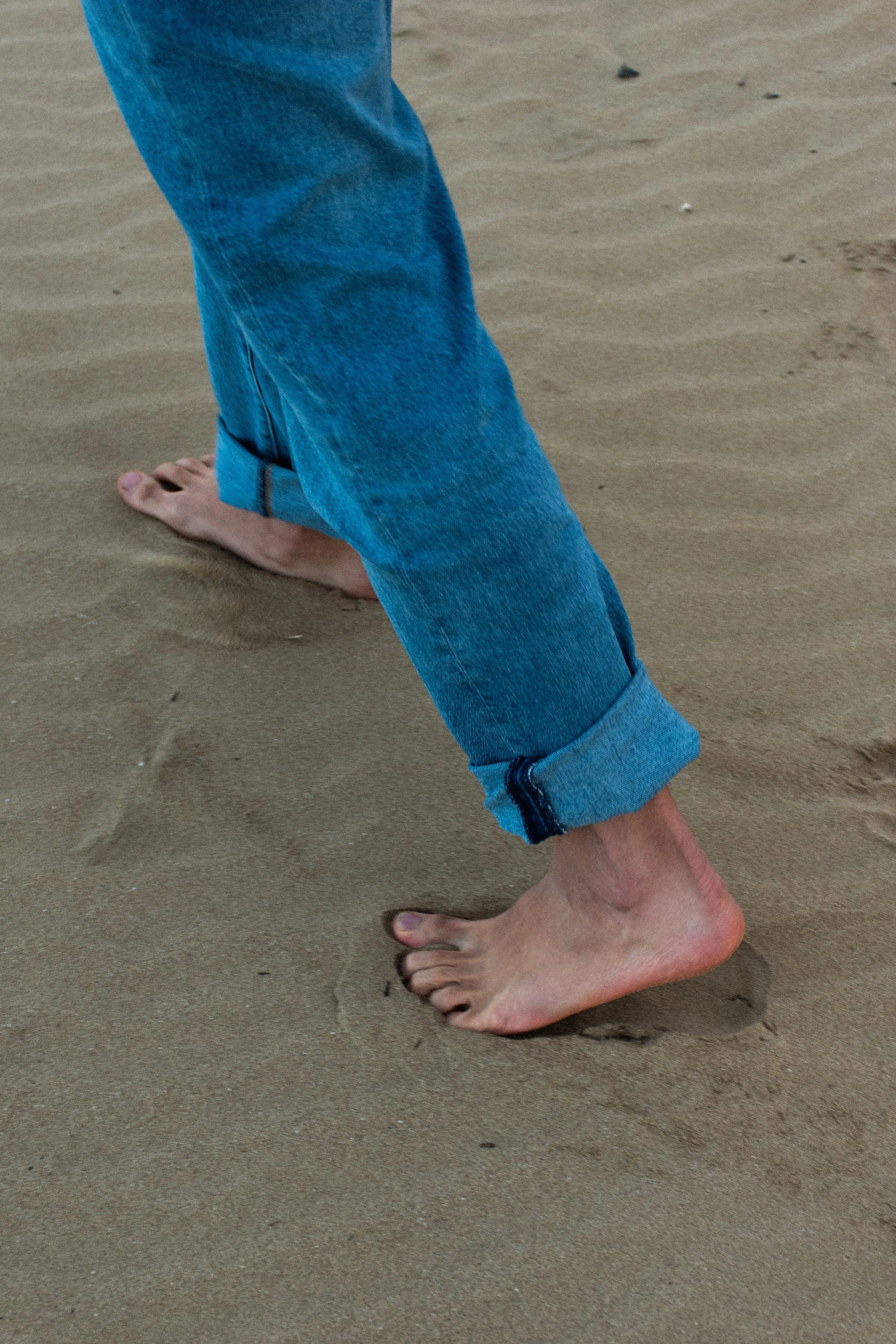 A person with rolled-up jeans barefoot walking or standing on the sand at the beach.