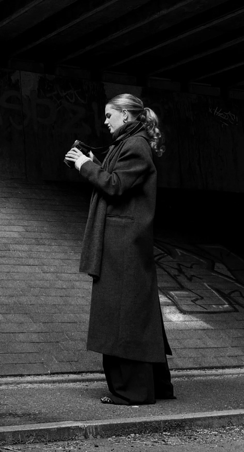 A woman wearing a long coat and a scarf, holding a camera, standing on a sidewalk in front of a graffiti-covered brick wall.