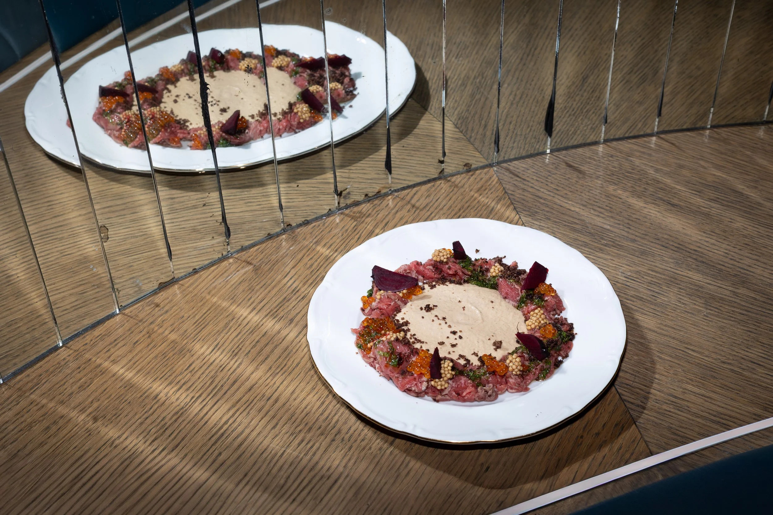 A white plate with raw steak tartare topped with a creamy sauce and garnished with herbs, vegetables, and sauces, viewed through a glass or mirror that reflects the plate.