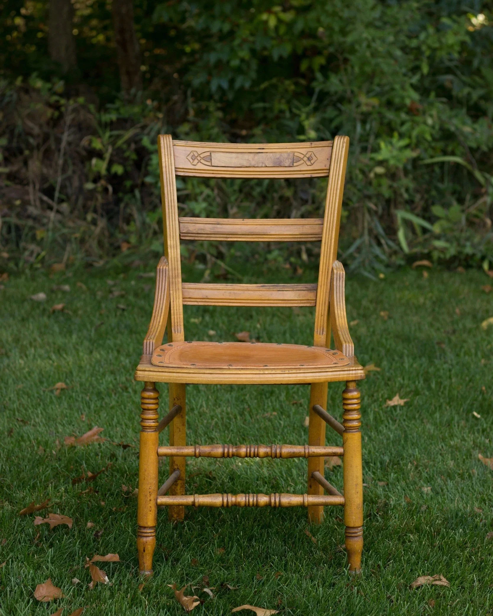A wooden chair with carved decorations on the top rail of the backrest, placed on a grassy field with fall leaves. A dense green hedge and trees are in the background.