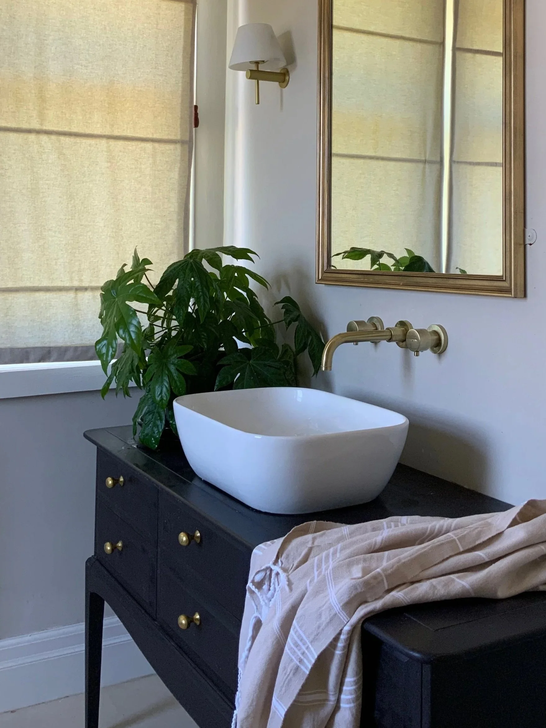 A black bathroom vanity with gold knobs, a white vessel sink, and a gold faucet underneath a window with beige shades. There is a potted green plant on the vanity, a gold-framed mirror on the wall, a wall-mounted lamp, and a beige striped towel.
