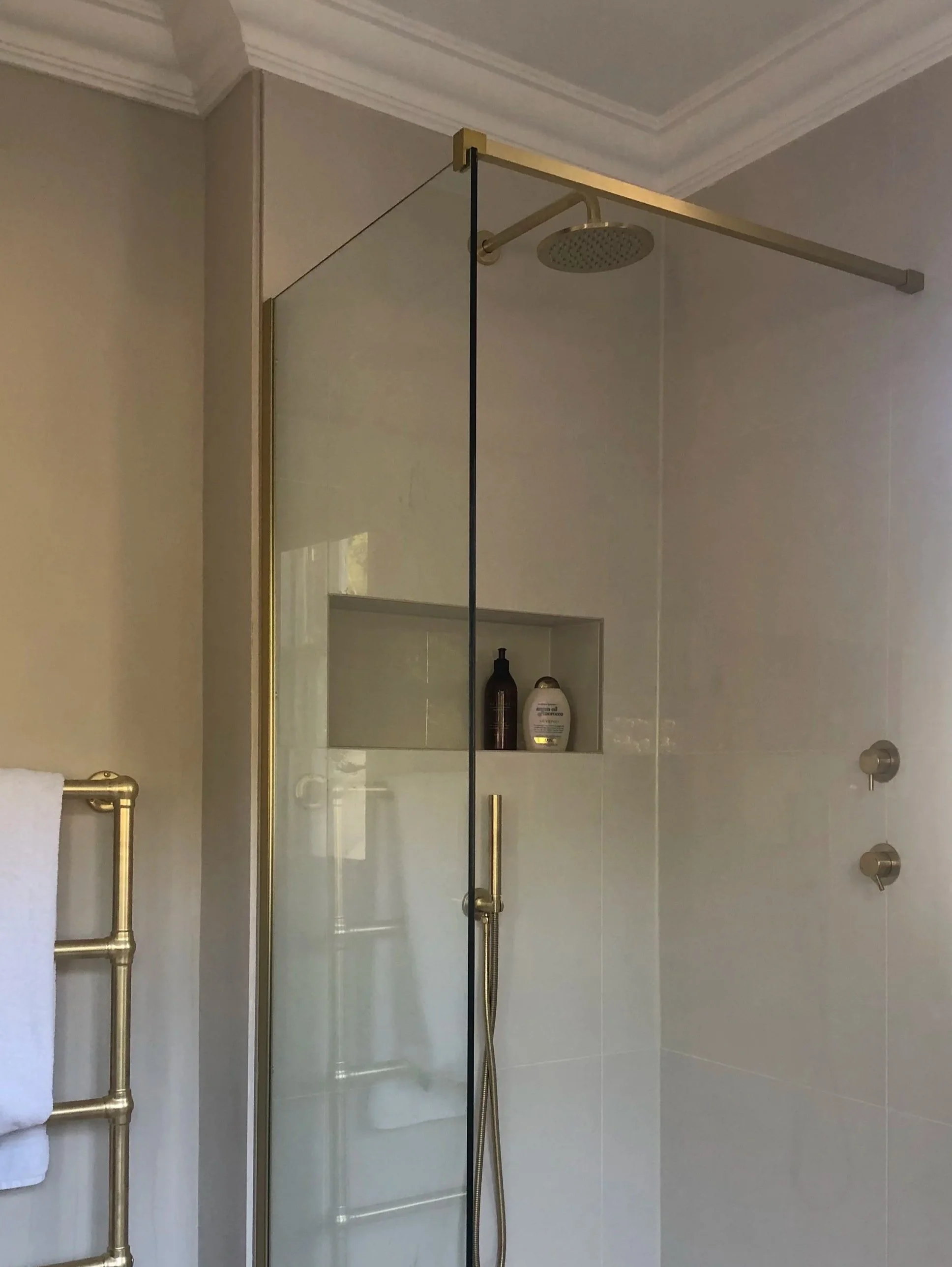 A walk-in shower with a glass door, a ceiling-mounted showerhead, and built-in wall shelves holding shampoo and body wash bottles. Gold-colored wall fixtures and a white towel hanging on a gold towel bar are visible.