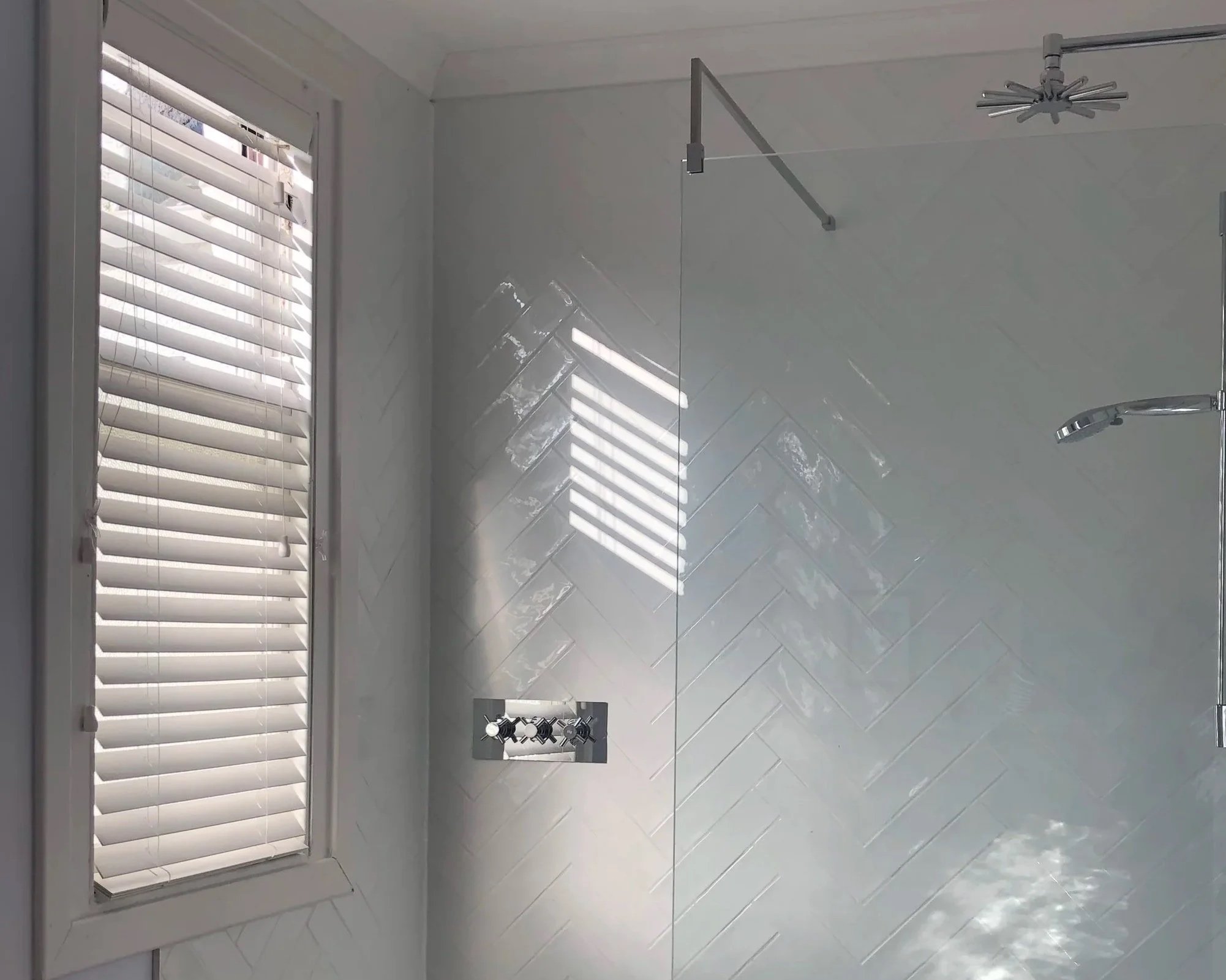 A bathroom with white tiled walls, a window with white horizontal blinds, a modern chrome shower head, and a square shower control with two knobs, all illuminated by natural light.