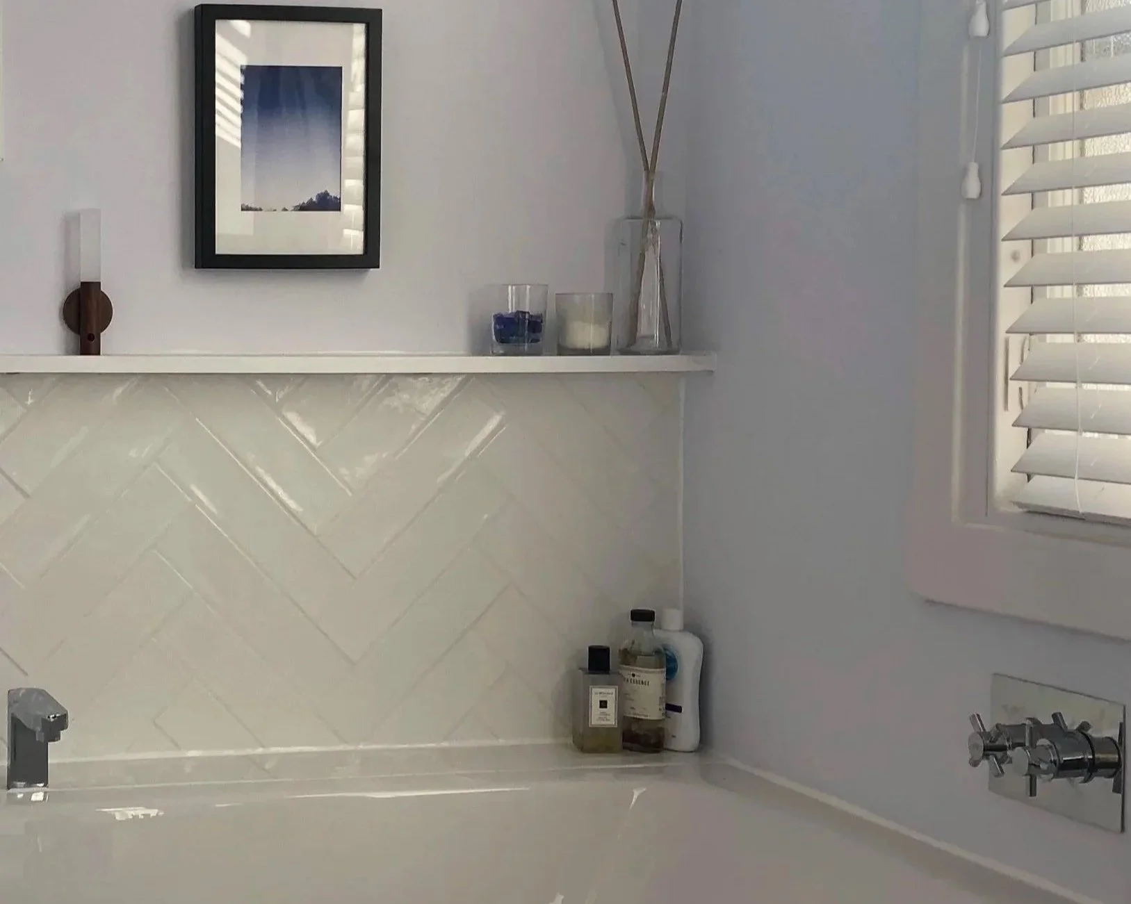 A bathroom corner with a white tiled wall, a window with white blinds, and a white shelf above the tiles holding decorative items including a framed picture, candles, and a clear vase with sticks. There are toiletries and bottles on the counter below