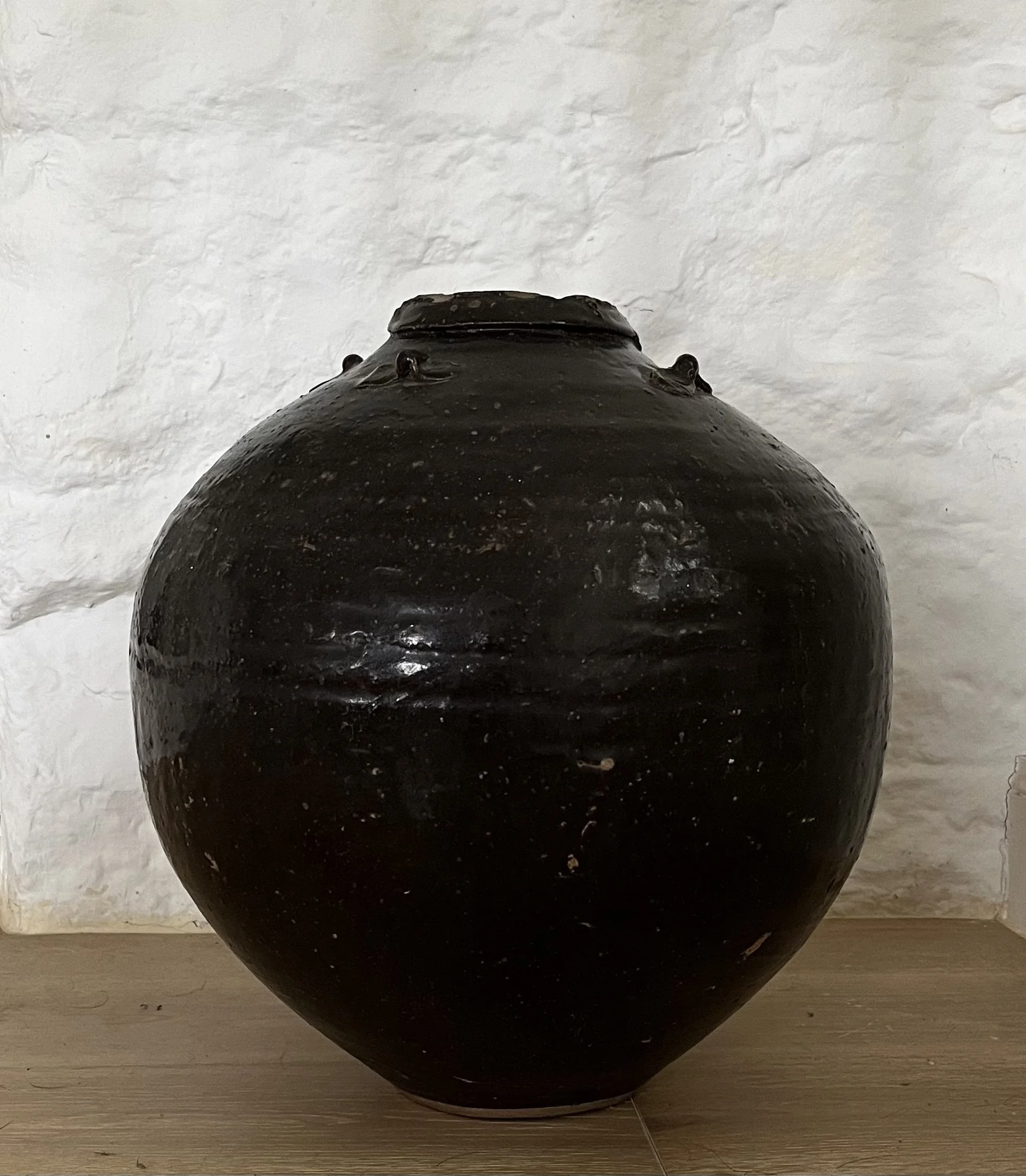 Close-up of a black clay pot with a rounded shape, set against a white textured wall and wooden floor.