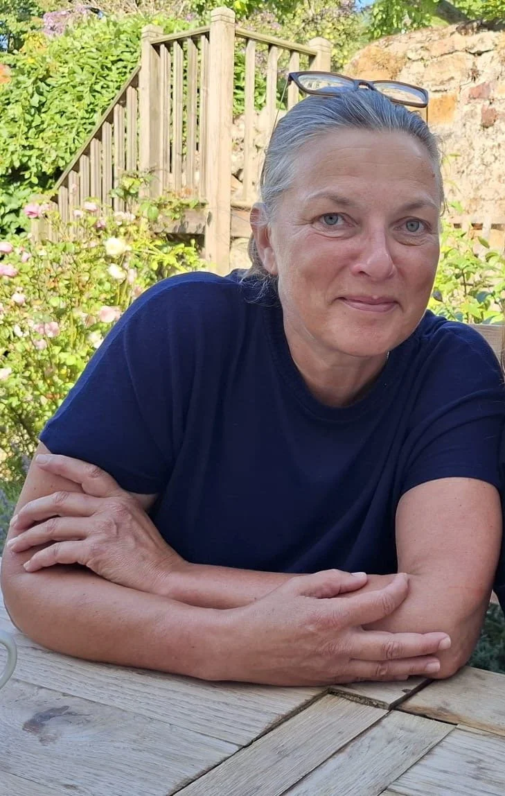 Sarah sitting outdoors at a wooden table, with a garden and wooden fence in the background, wearing a blue t-shirt
