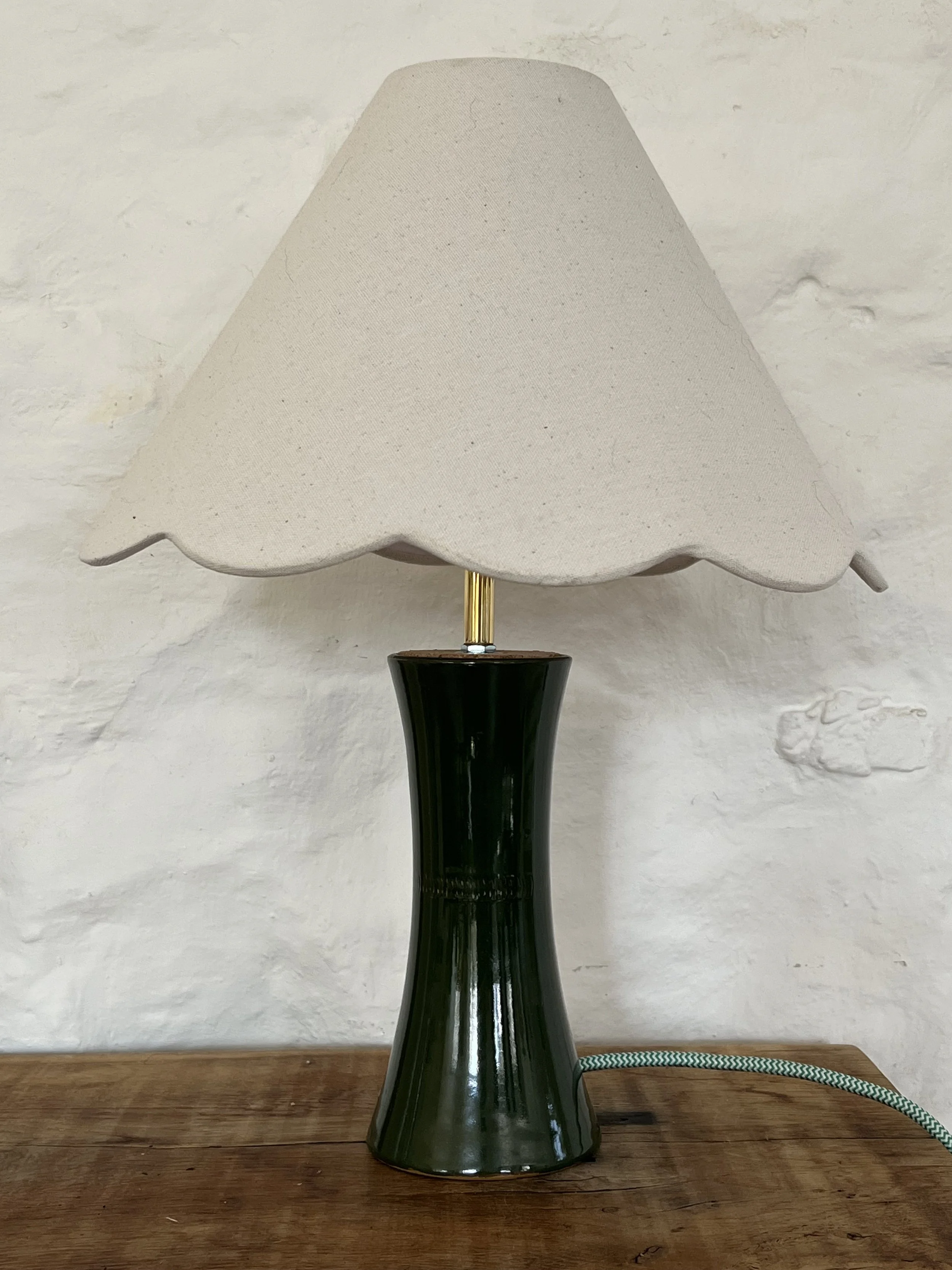 A modern table lamp with a black base and a large, textured white fabric shade on a wooden surface against a white brick wall.