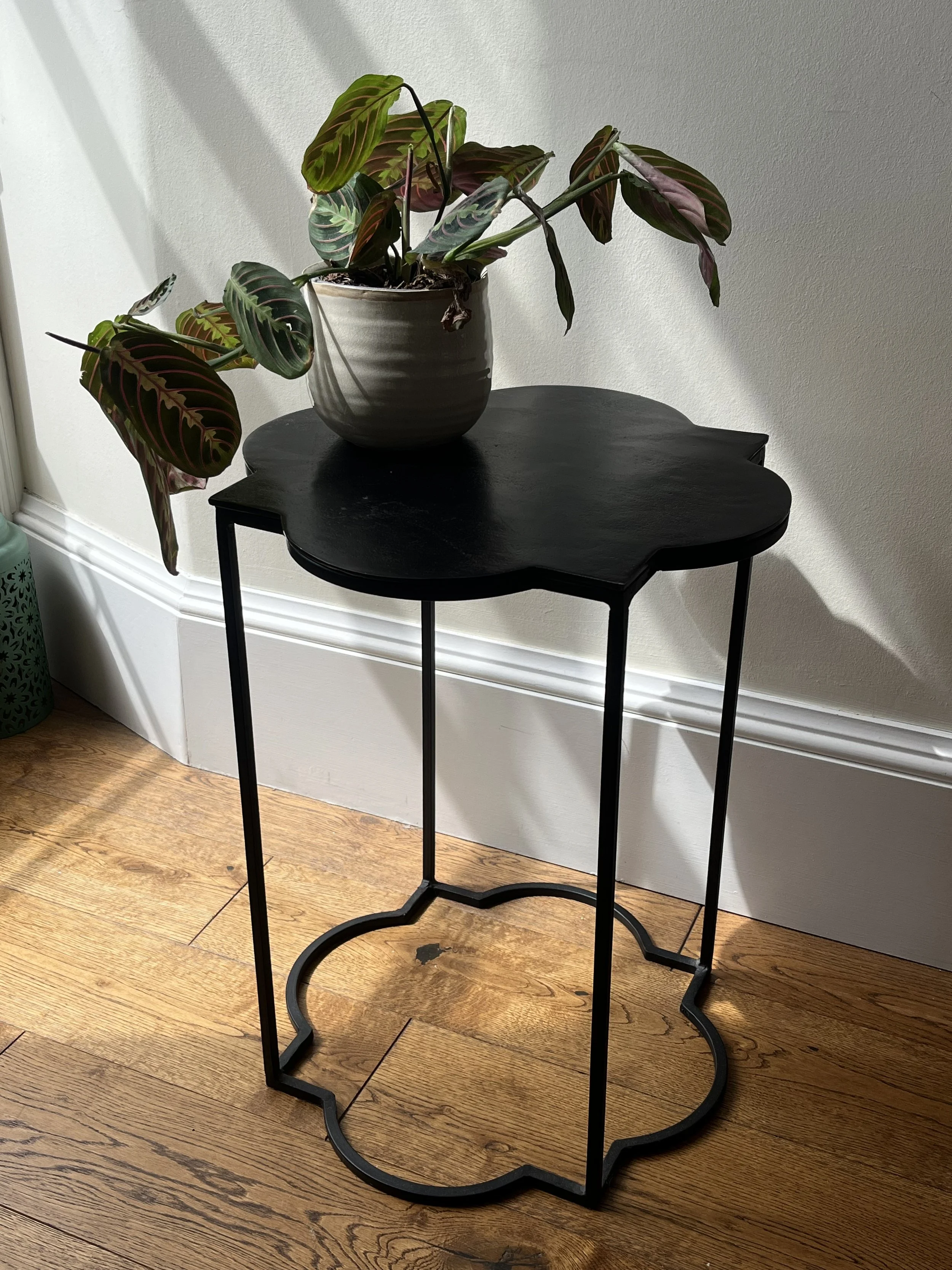 A black metal side table with a potted plant on top, next to a white wall and hardwood floor.