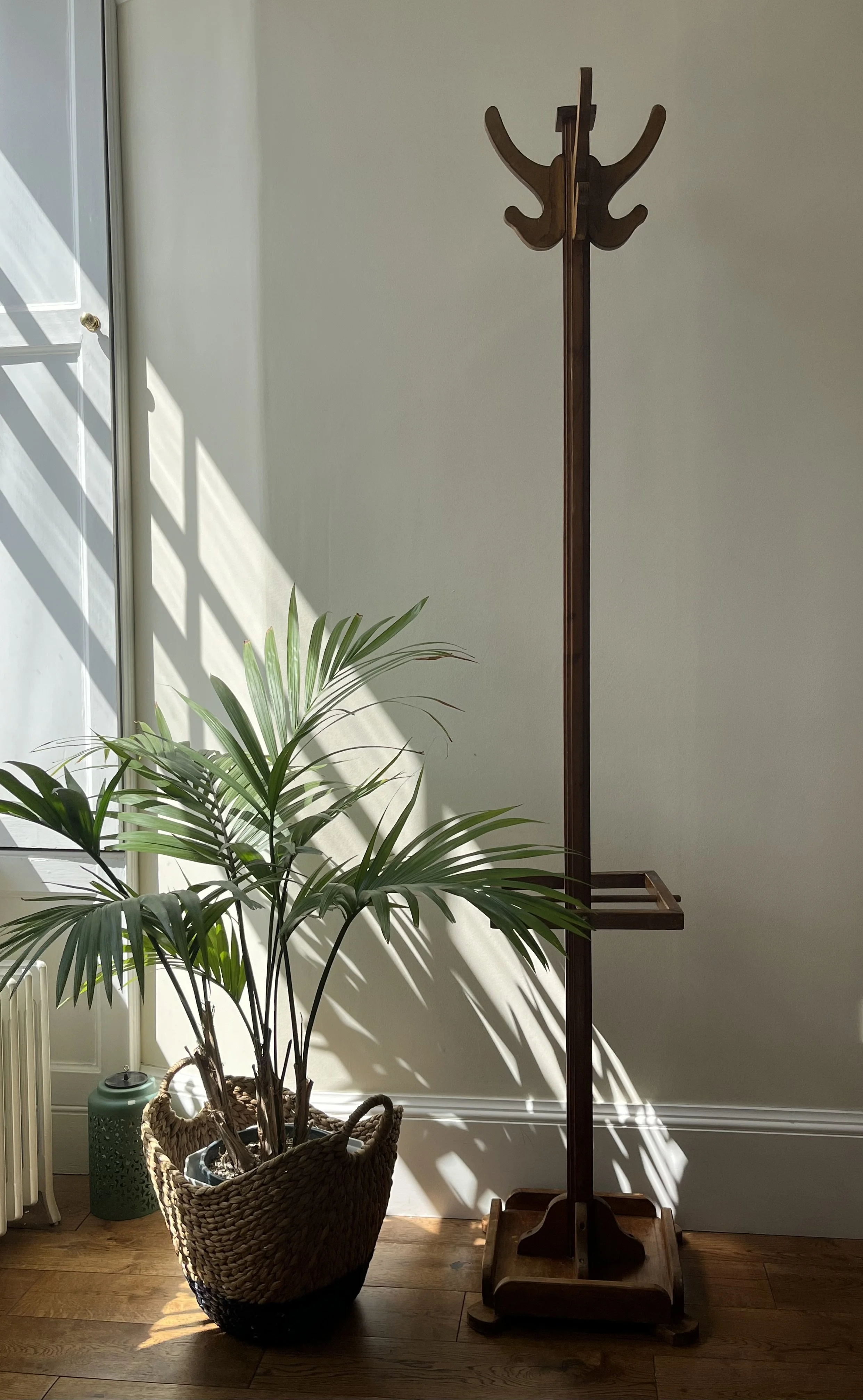 A potted palm plant in a woven basket, next to a wooden coat stand with hooks and a small shelf, near a window with sunlight streaming in.