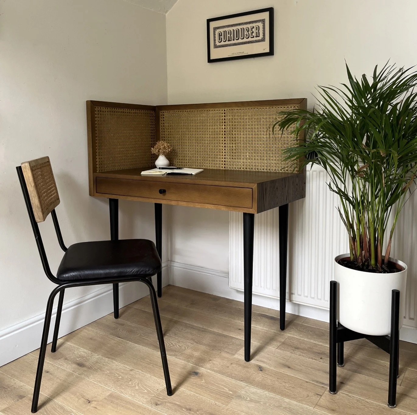 A small wooden desk with black metal legs, a drawer, and a woven rattan back panel, placed in a corner of a room. On the desk are an open book, a white vase with dried flowers, and a closed notebook. Next to the desk is a black chair with a wooden ba
