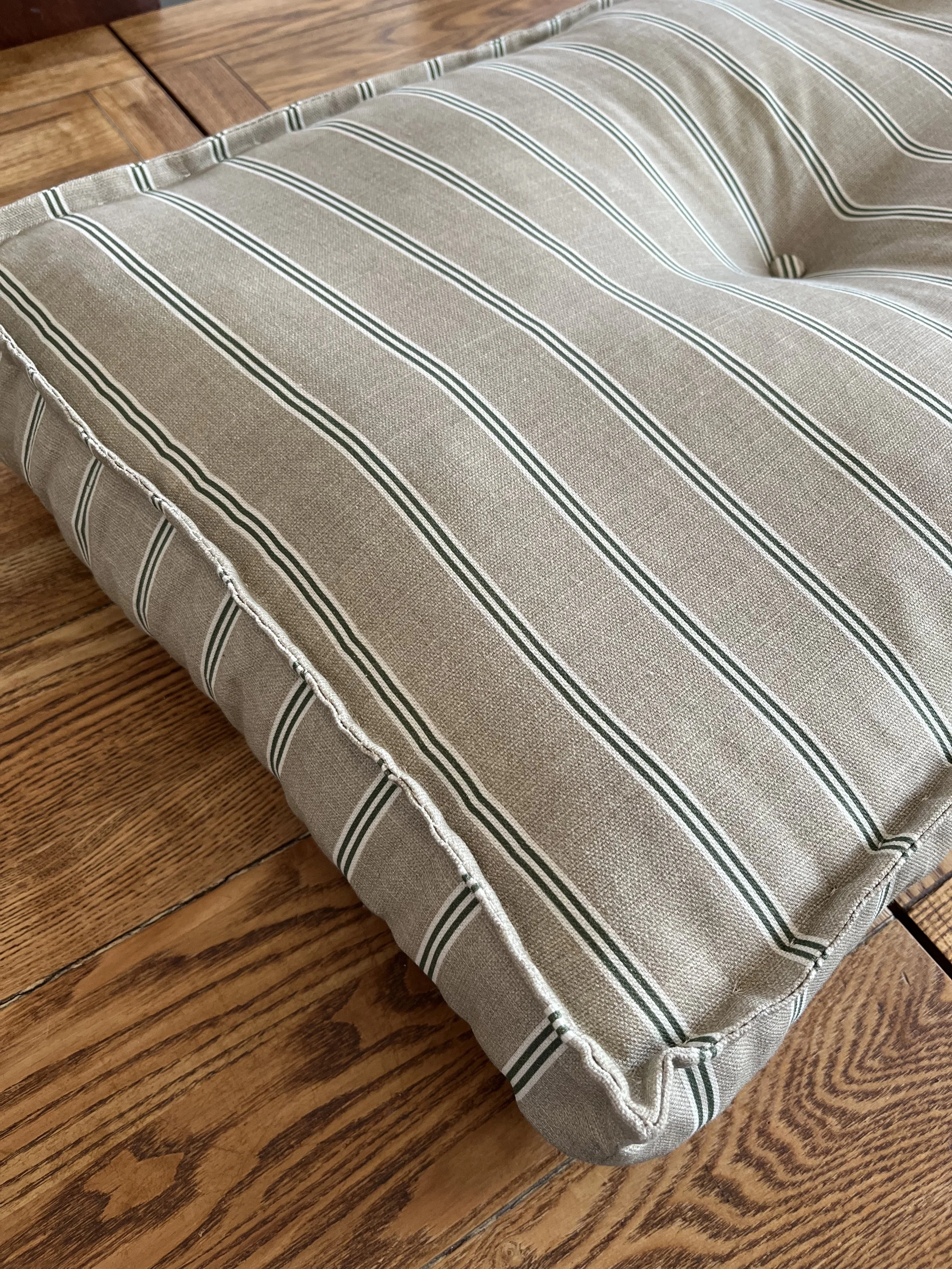 A beige fabric cushion with green and white stripes placed on a wooden floor.