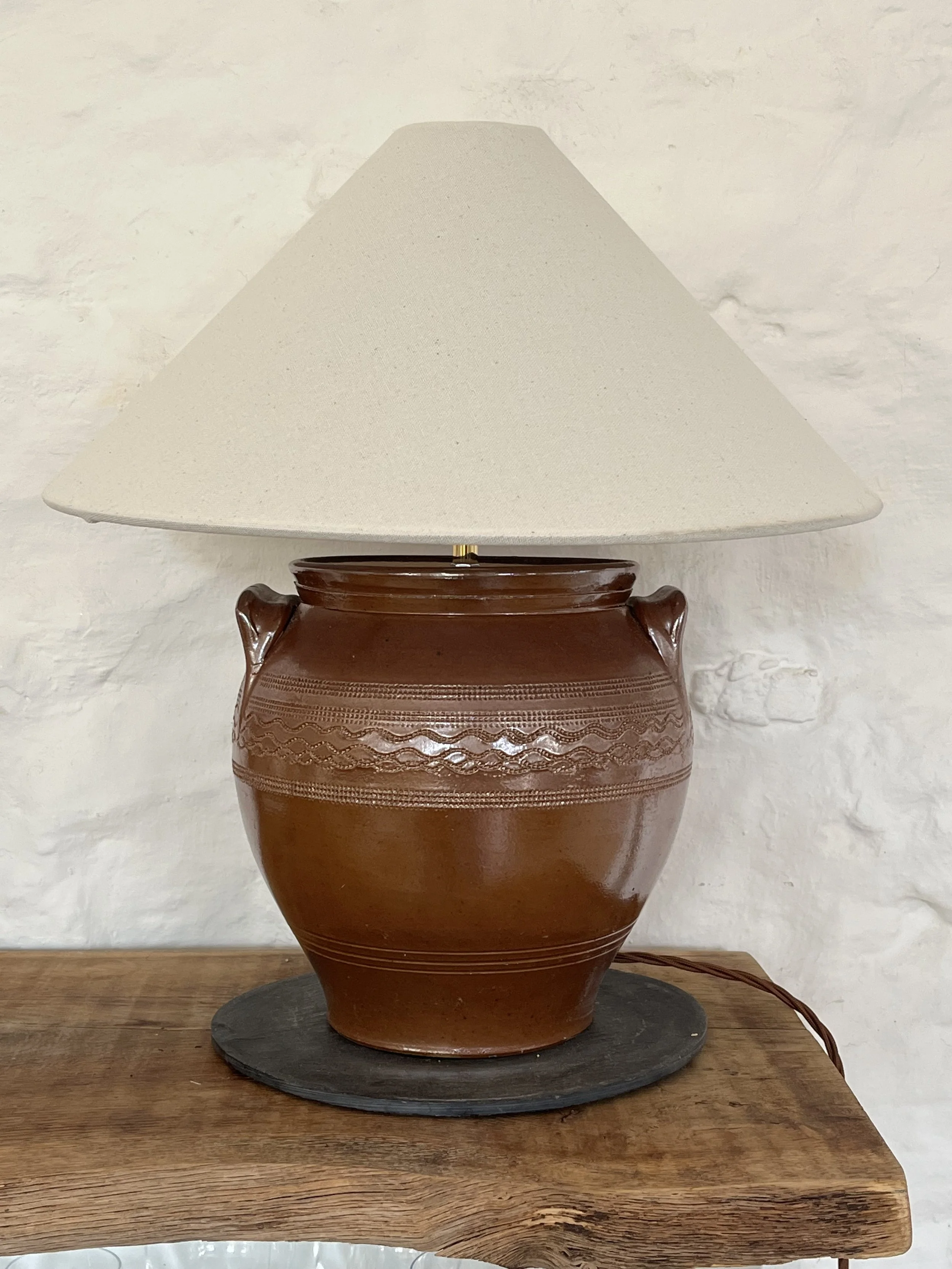 A large decorative ceramic vase with a carved design, sitting on a black circular tray on a wooden table, with a white textured wall in the background.