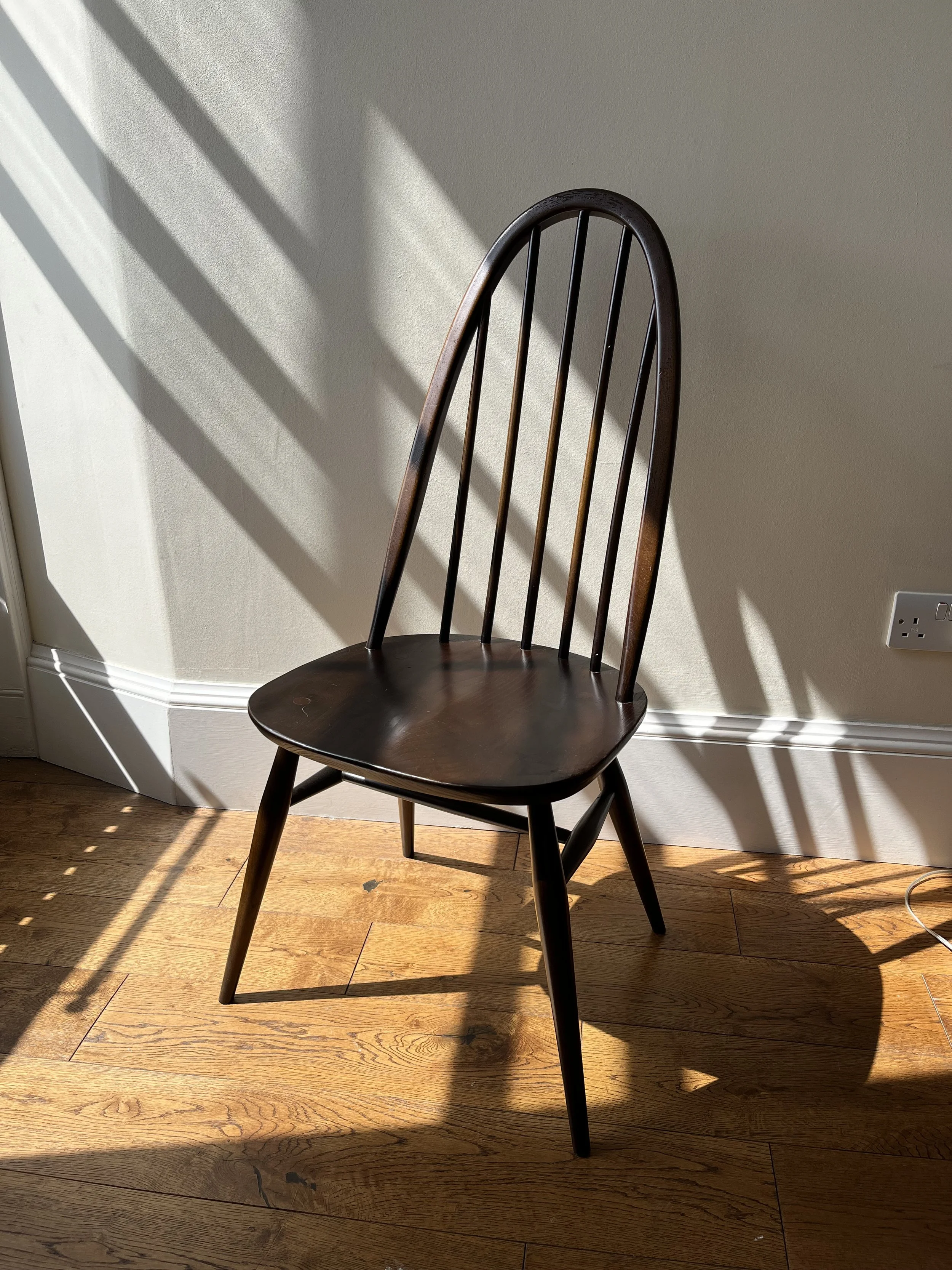 A wooden chair with a high arched back and vertical slats, placed on a hardwood floor near a wall with sunlight and shadows.