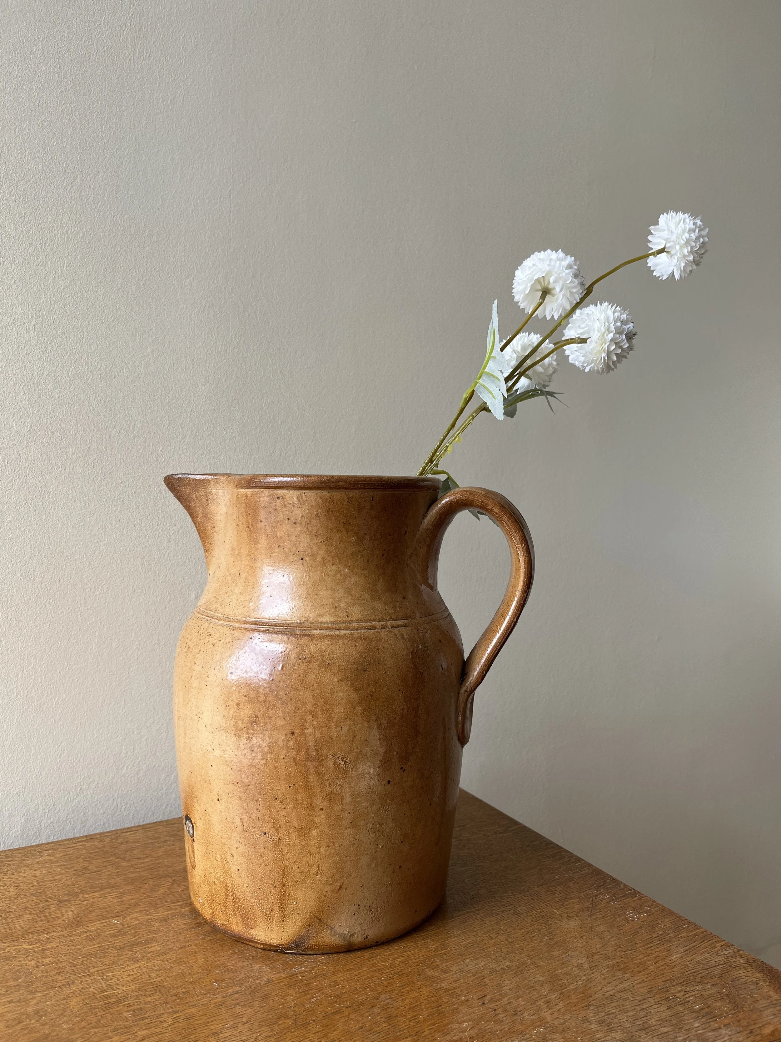 A brown ceramic vase with a handle on the right, containing white artificial flowers with long stems, placed on a wooden surface against a plain beige wall.