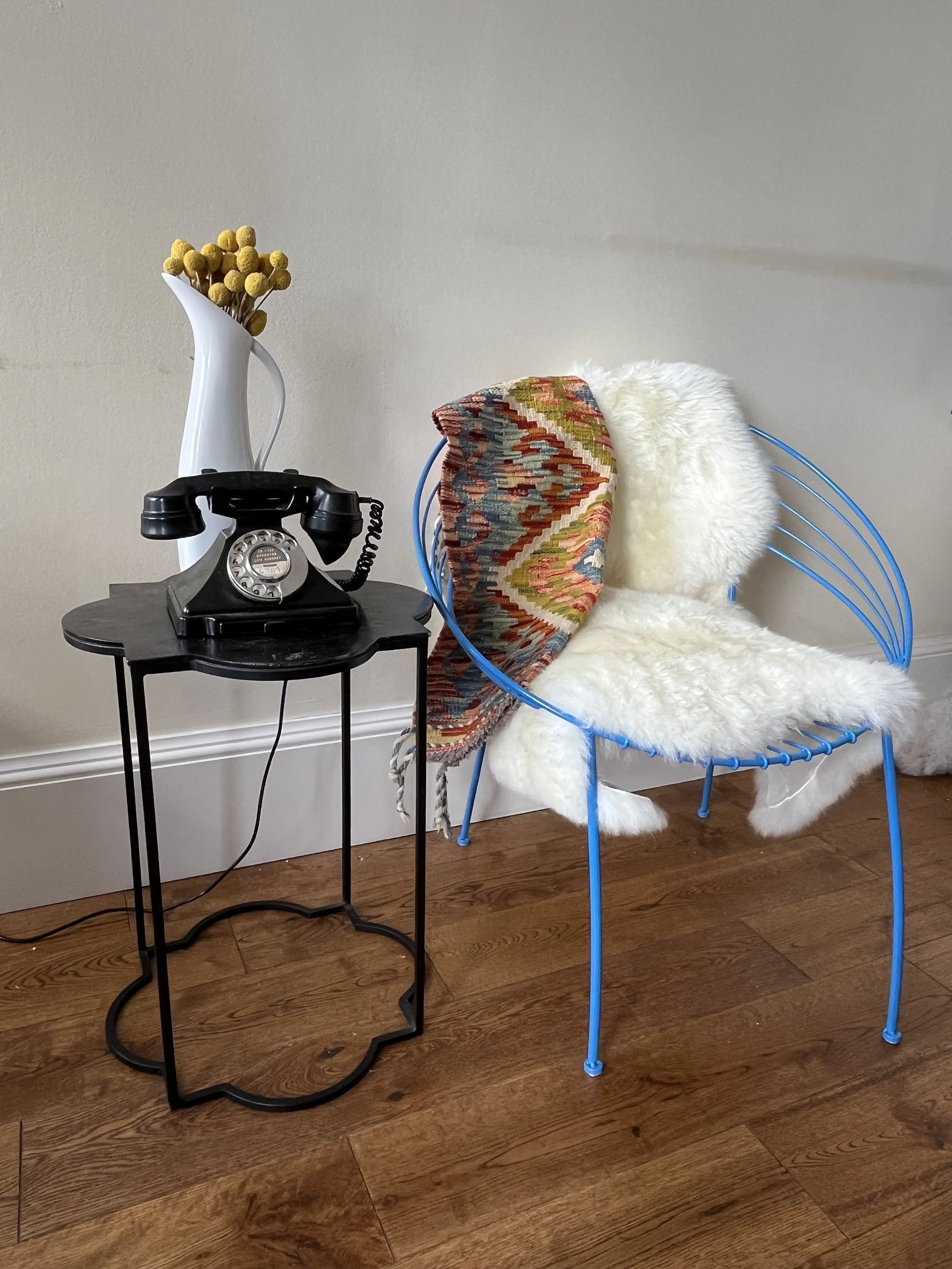 A round black side table with an antique black rotary phone on top. Behind the table, there is a modern blue wireframe chair with a white fluffy throw and a colorful patterned fabric draped over the back. On the table, there is a white ceramic vase w